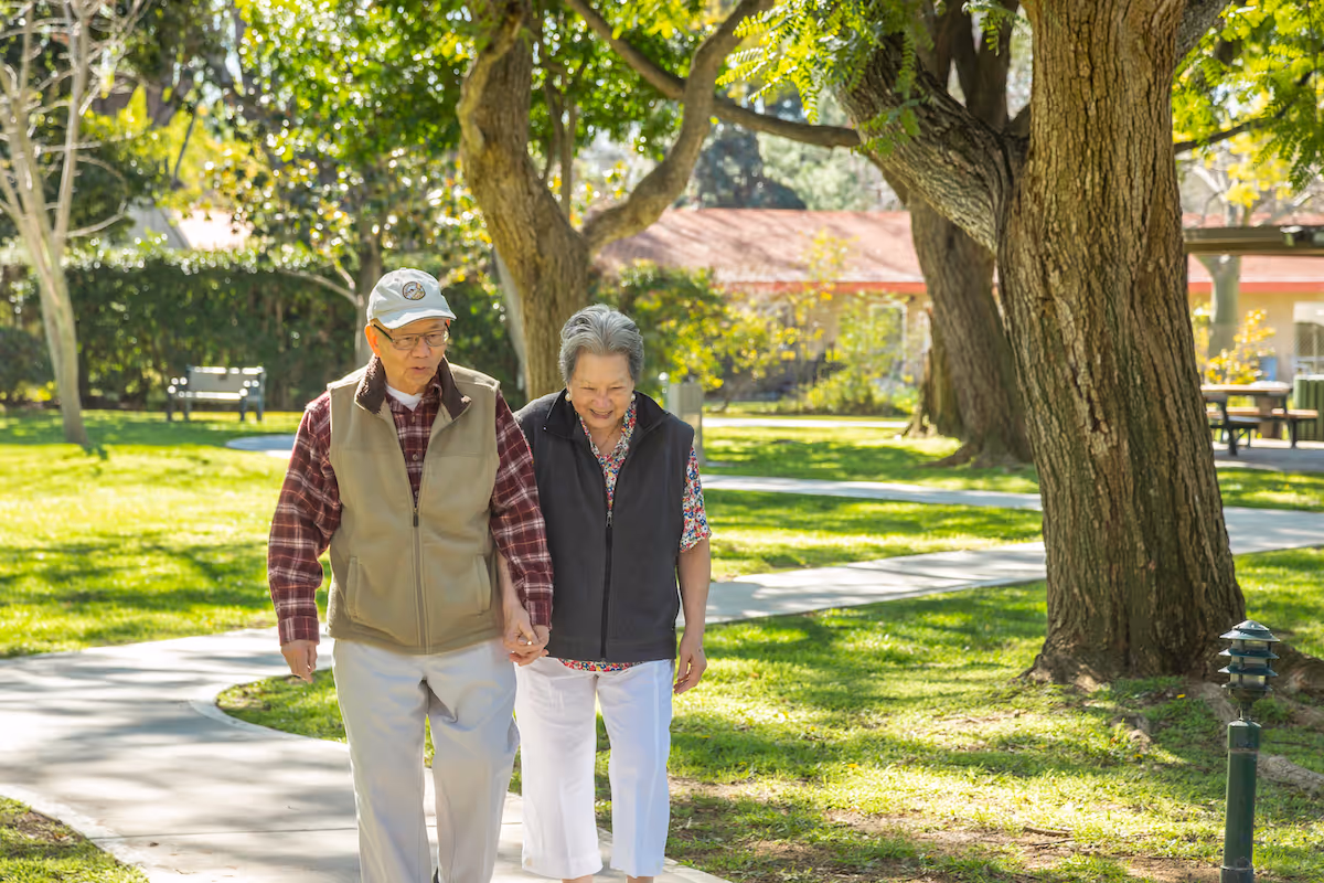 An elderly couple walking hand in hand on a paved path in a sunny garden area with large trees and green grass. The man is wearing a beige vest, plaid shirt, white pants, and a cap, while the woman is wearing a black vest over a floral shirt and white pants. There are benches and a building with a red roof in the background.