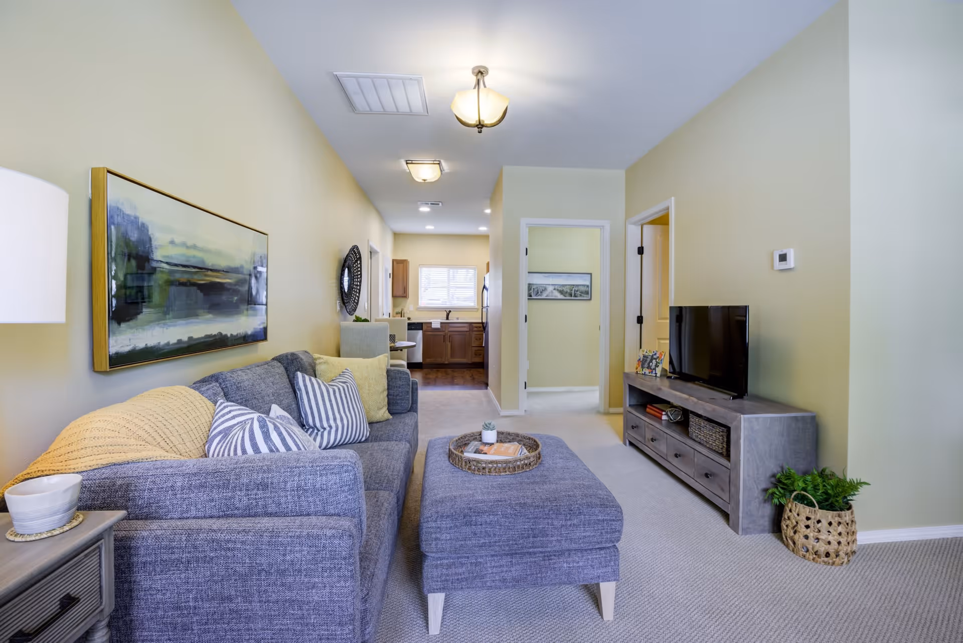 A cozy living room with a gray sofa adorned with striped and solid pillows, a matching ottoman with a tray on top, a wooden TV stand with a flat-screen TV, and a small potted plant in a woven basket on the floor. The room has light yellow walls, beige carpet, and ceiling lights. In the background, there is a view into a kitchen area with wooden cabinets and a window, as well as doorways leading to other rooms.