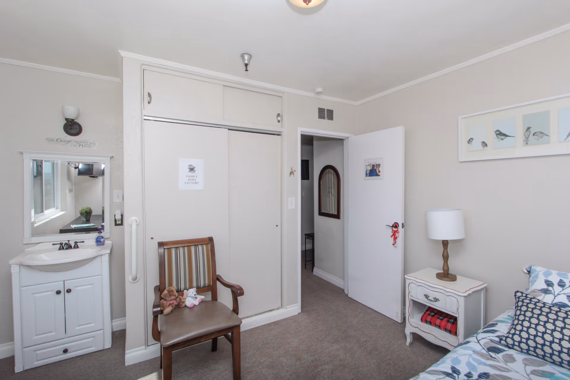 A cozy bedroom in a senior assisted living facility featuring a bed with patterned bedding and pillows, a small white nightstand with a lamp, a wooden chair with two stuffed animals on it, a closet with sliding doors, a small sink with a mirror above it, and a door leading to another room. The walls are painted light beige and decorated with framed bird artwork.