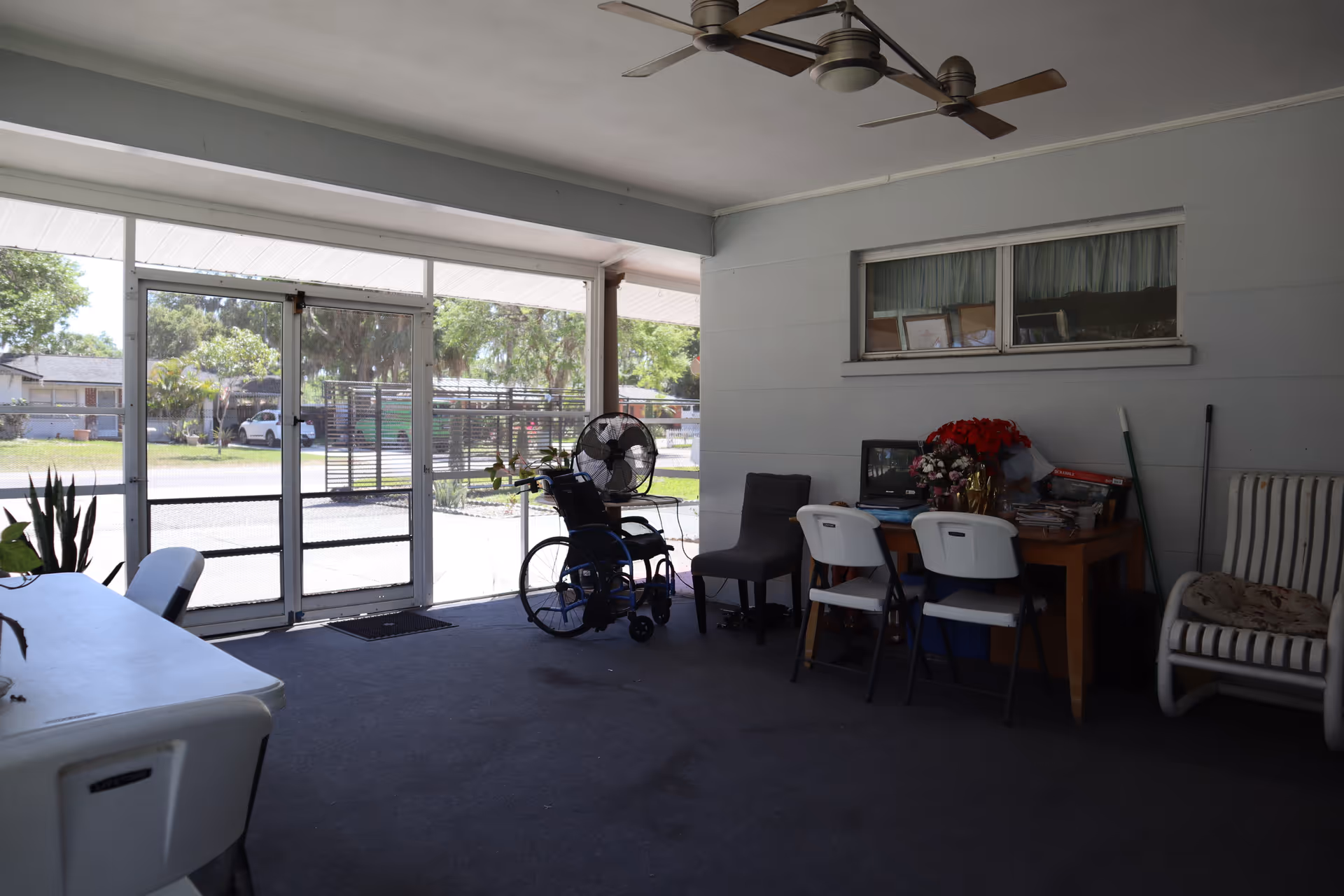 Enclosed screened porch with chairs, a wheelchair, a table with flowers and a ceiling fan, looking out to the street.