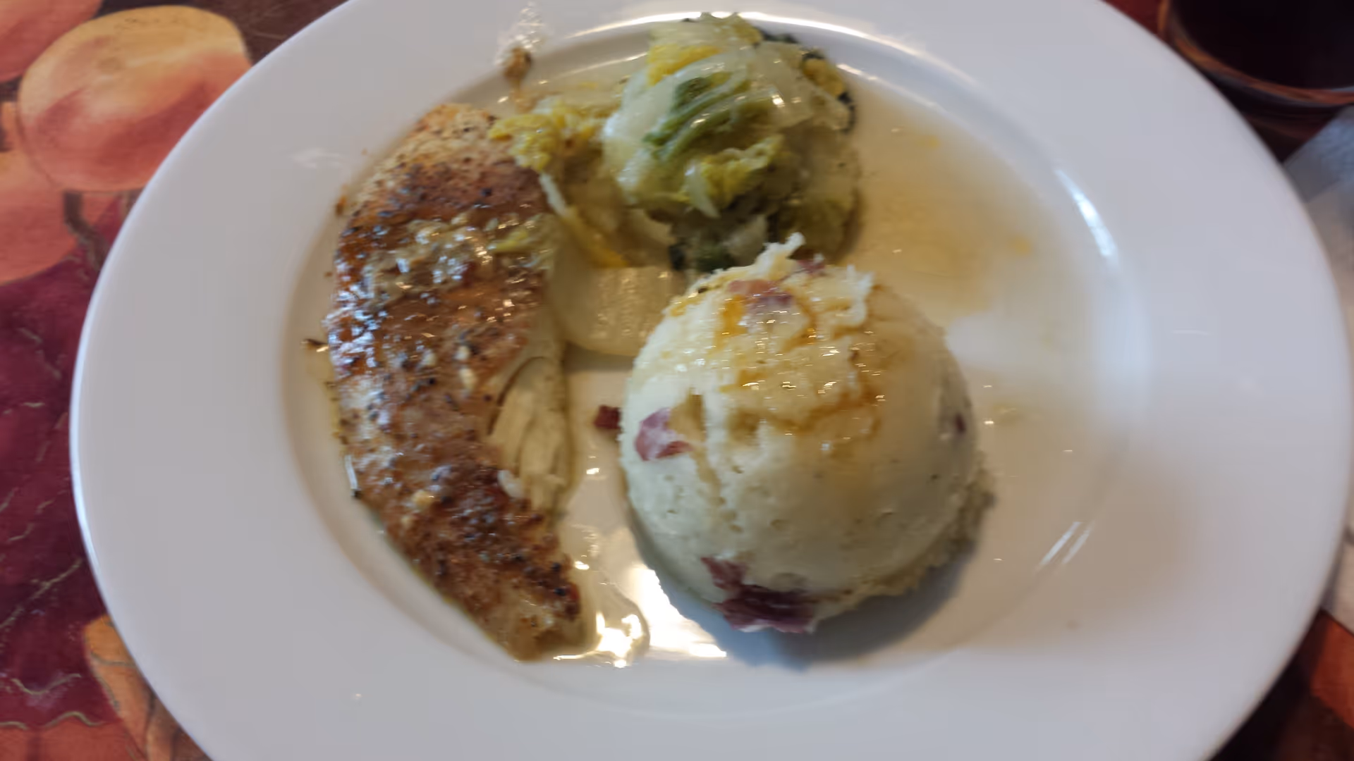 A white plate with a serving of seasoned cooked chicken breast, a scoop of mashed potatoes with bits of bacon, and a portion of cooked cabbage in a light sauce.