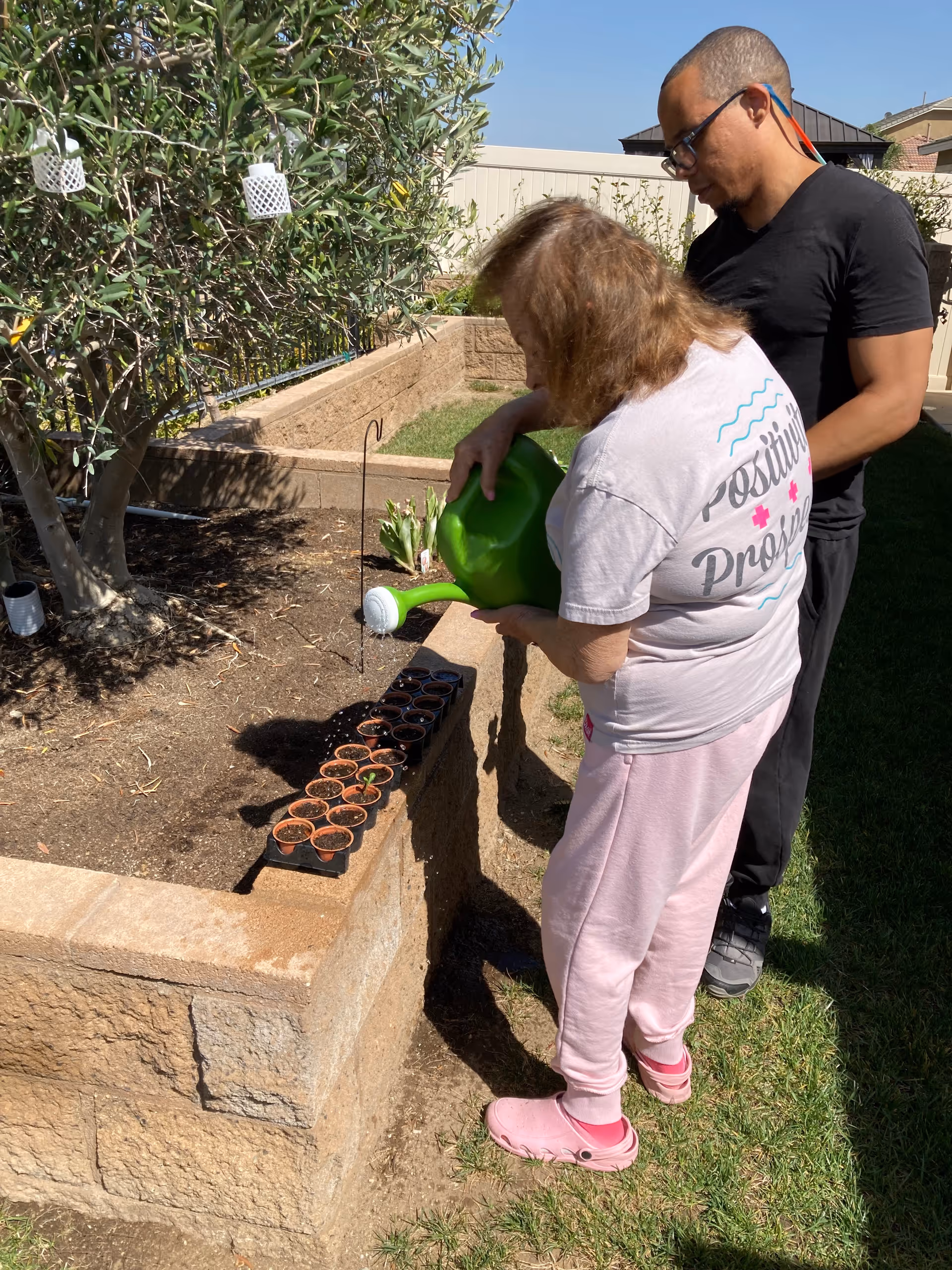 An elderly woman wearing a light pink outfit and pink shoes is watering small plants in pots placed on a raised garden bed. A man in a black shirt and pants stands beside her, watching. The garden bed is made of stone blocks and there is a tree and some greenery in the background under a clear blue sky.