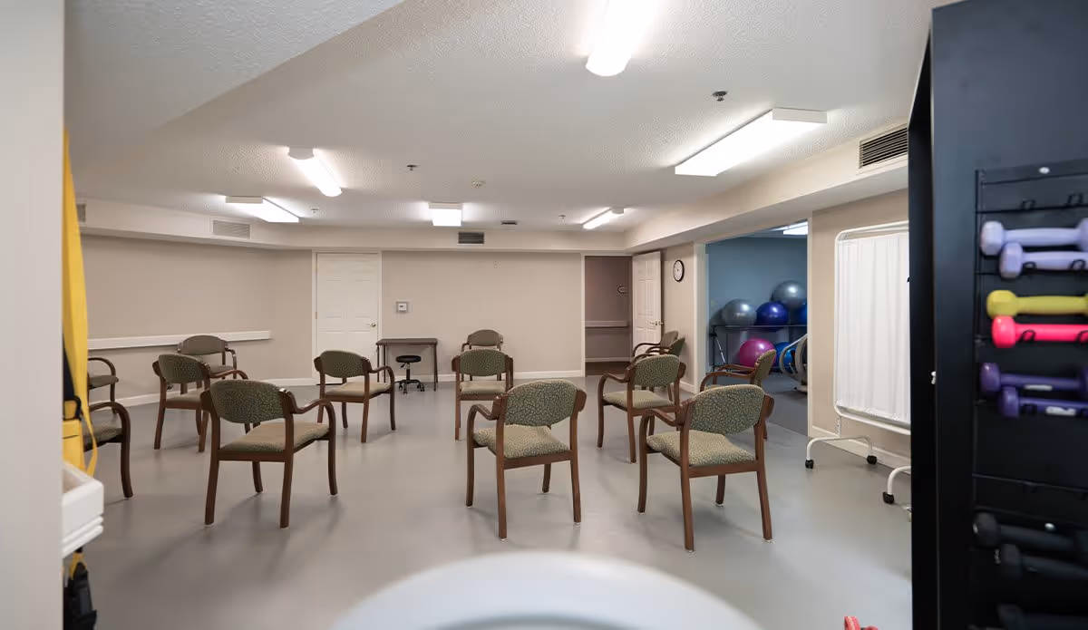A spacious room with several green cushioned chairs arranged in a semi-circle on a gray floor. The room has beige walls and multiple fluorescent ceiling lights. In the background, there is an open doorway leading to another room with exercise balls and fitness equipment. A rack with colorful dumbbells is visible on the right side of the image.