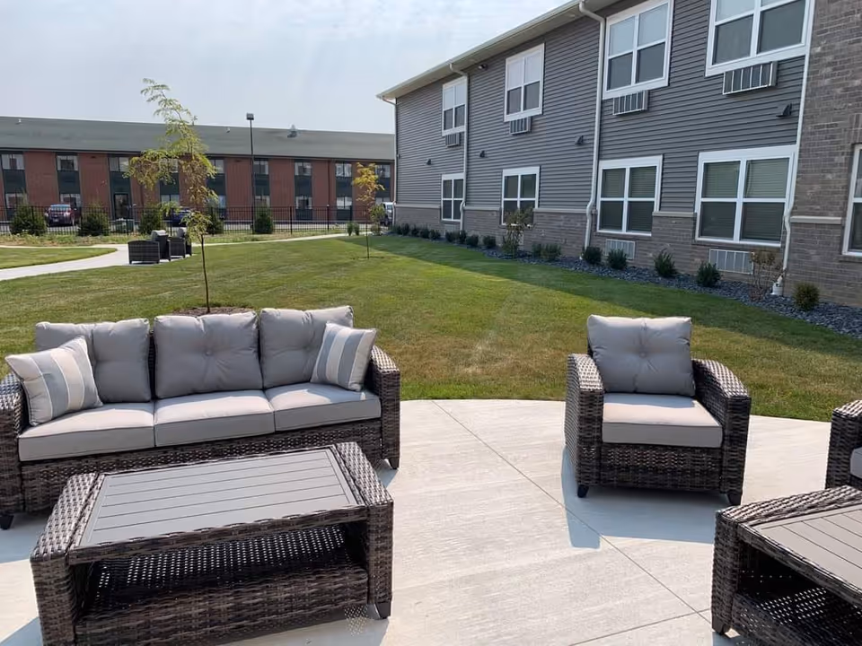 Outdoor patio area with cushioned wicker furniture including a sofa, armchairs, and a coffee table on a concrete surface. The background shows a grassy lawn, young trees, and a two-story building with multiple windows and air conditioning units.