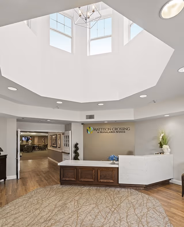 Reception area of Mattison Crossing at Manalapan Avenue featuring a white and wood front desk, a decorative plant, a vase with flowers, a large circular patterned rug, and a ceiling with a geometric skylight letting in natural light.