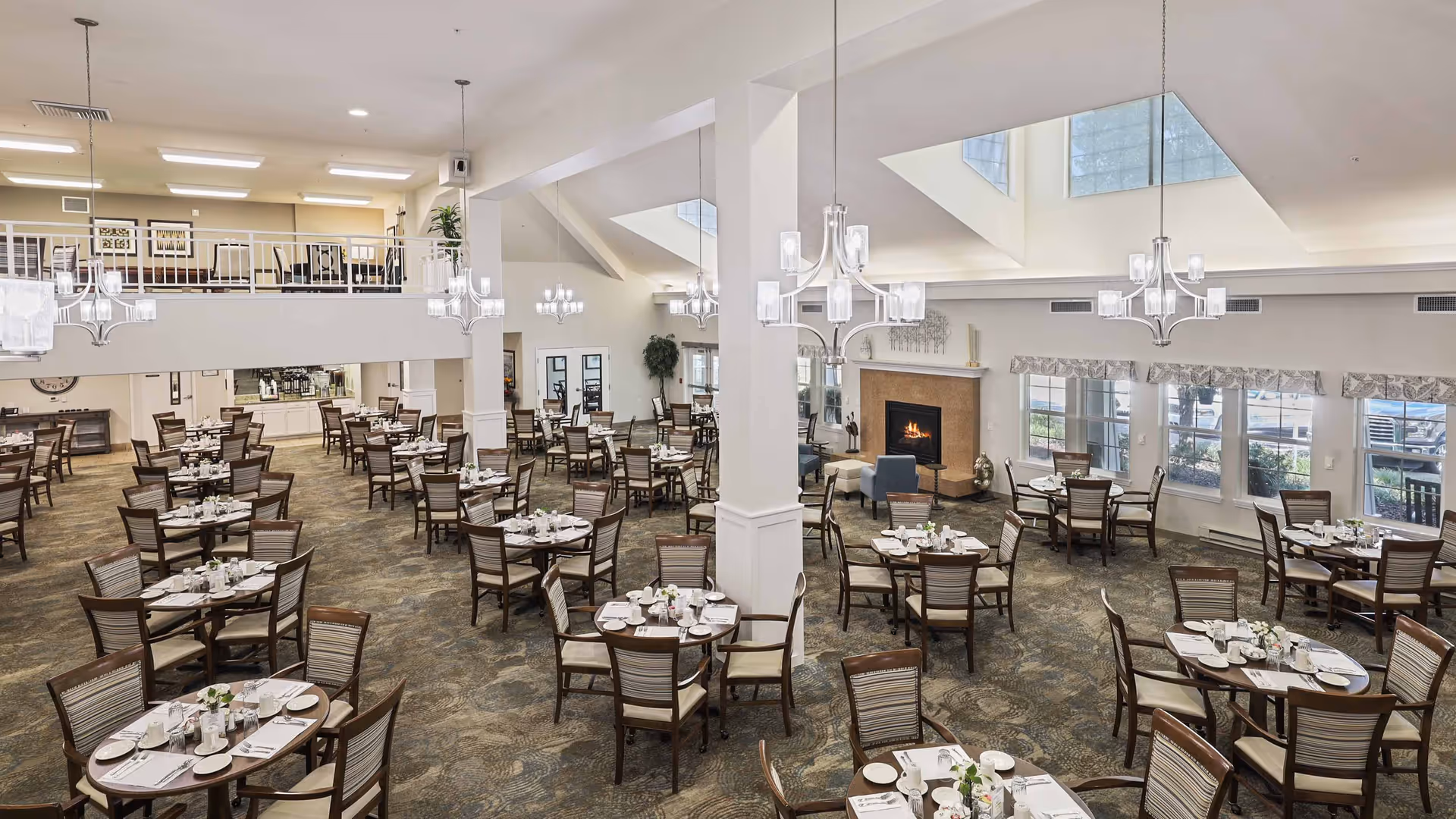 Spacious dining room with multiple round and rectangular tables set with white plates, cups, and silverware. The room features high ceilings with large windows allowing natural light, modern chandeliers, a fireplace with seating, and a mezzanine level overlooking the area.