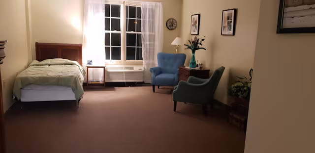 A spacious studio-style bedroom with a single bed by a window, two upholstered chairs, side tables and soft lighting.