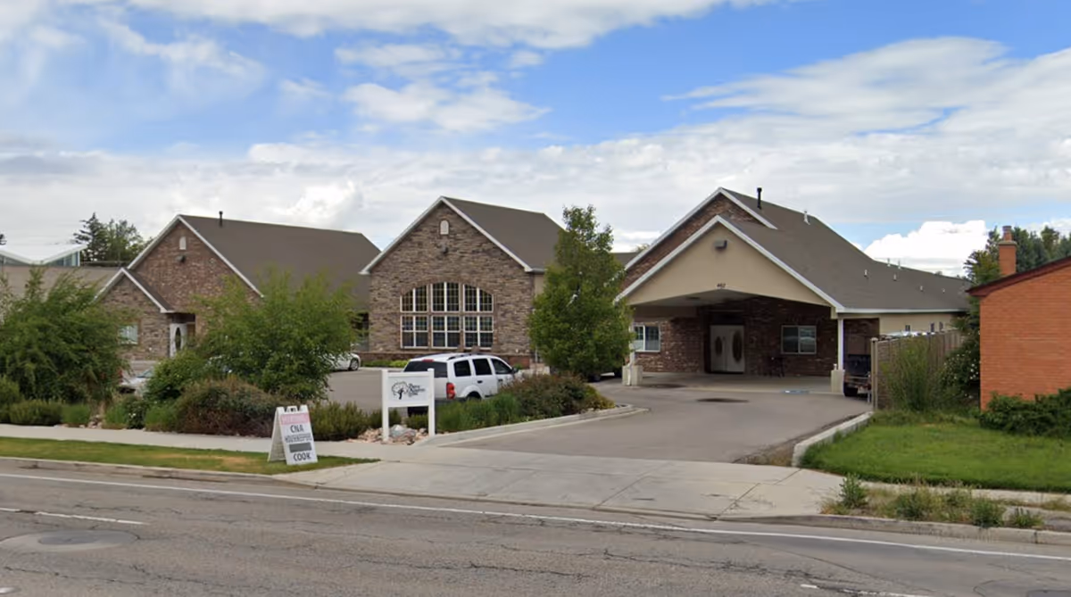 Exterior view of Rocky Mountain Care - River Pointe Assisted Living facility showing a large brick building with multiple peaked roofs, a covered entrance, several windows, and a parking area with cars. There are trees and shrubs around the building and a sidewalk along the street in front.