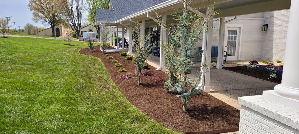 A well-maintained outdoor garden area beside a white building with a covered porch supported by white columns. The garden features neatly mulched beds with small trees and shrubs, bordered by a green lawn under a clear sky.
