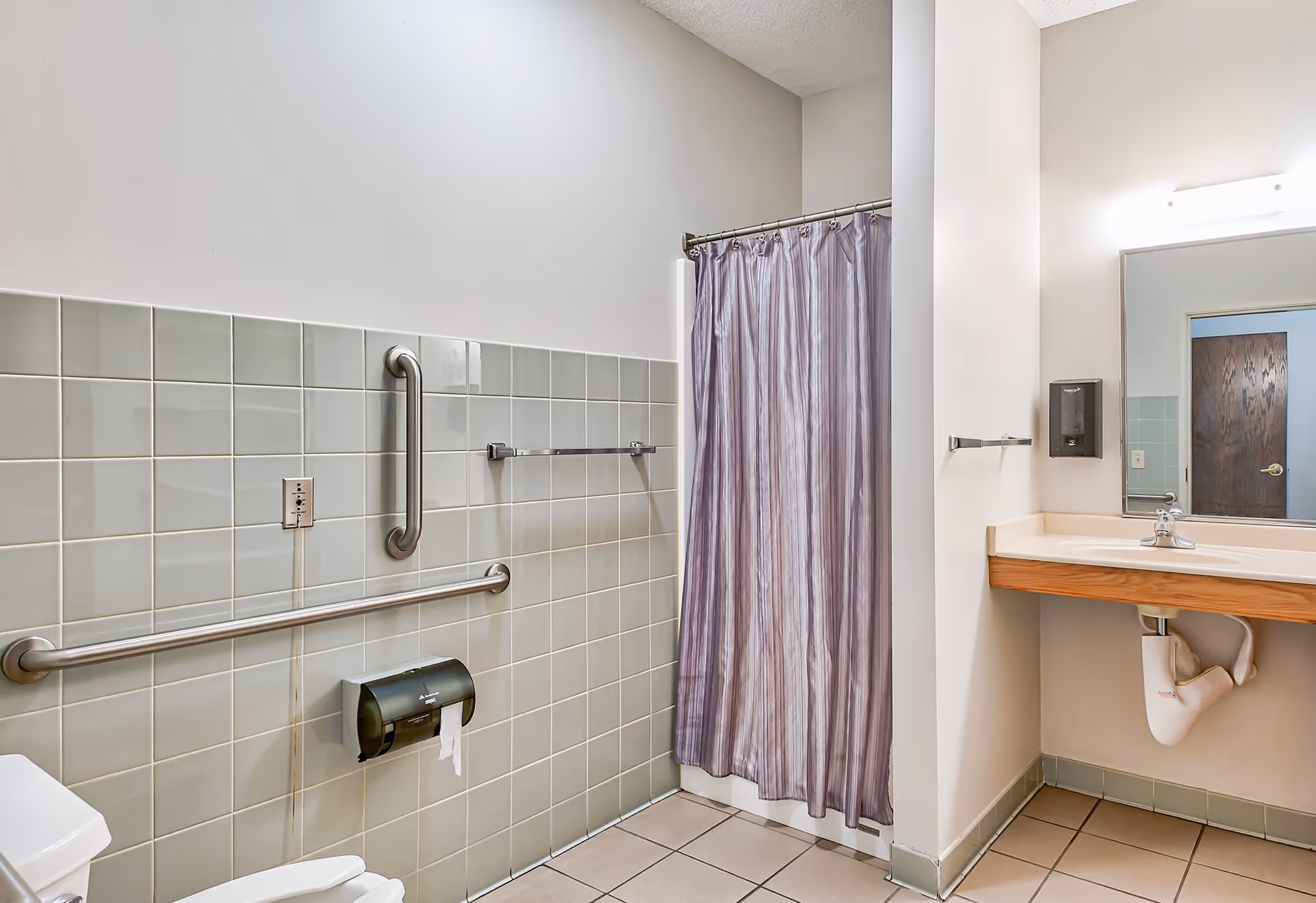 A clean and accessible bathroom with light gray tiled walls and beige floor tiles. The bathroom features a toilet with grab bars on the wall, a shower area with a purple shower curtain, and a sink with a large mirror above it. There is a soap dispenser mounted on the wall next to the mirror.