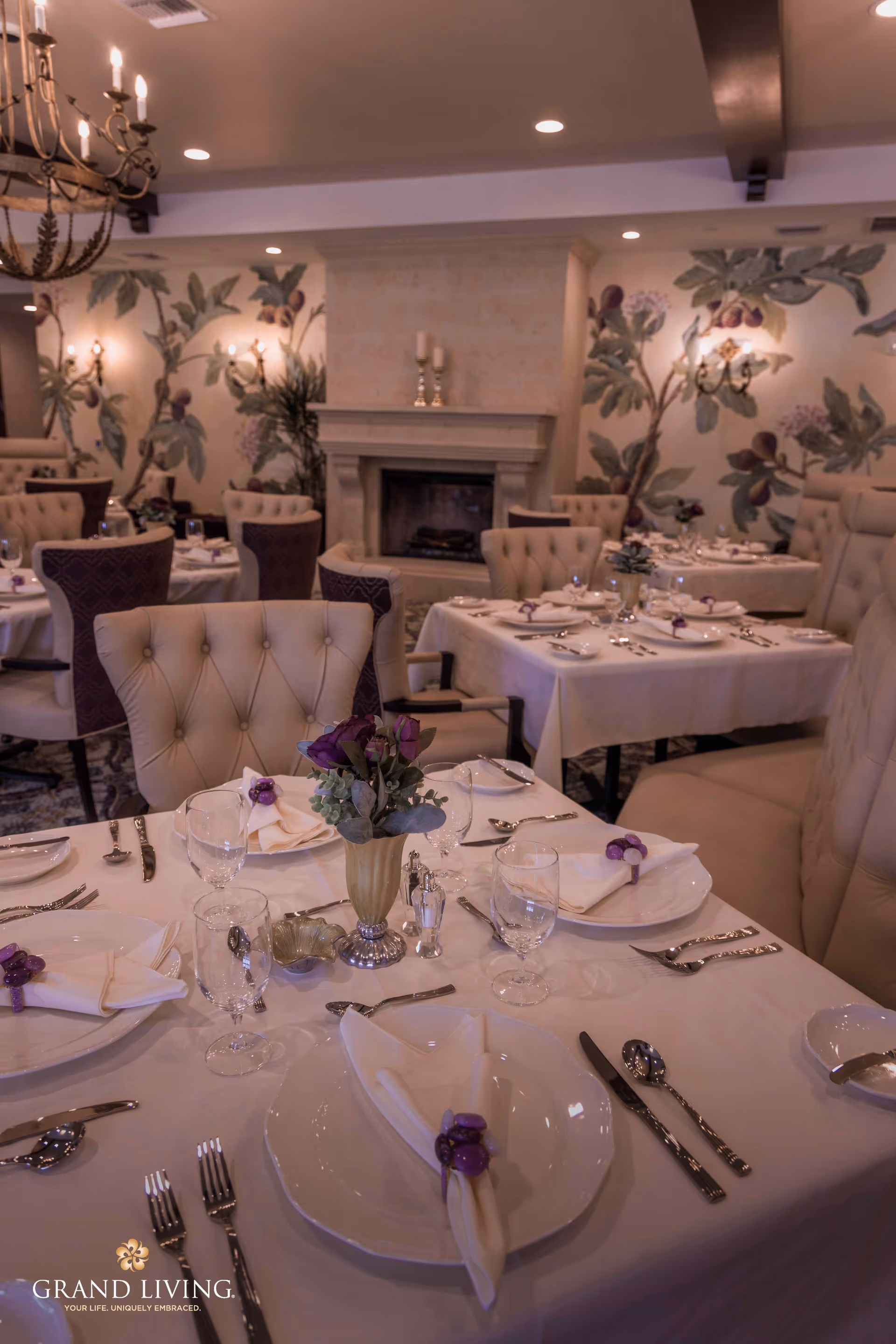 Elegant dining room with tables set with white tablecloths, plates, silverware, glassware, and napkins adorned with purple napkin rings. The room features tufted chairs, a fireplace, floral wallpaper, and soft lighting from chandeliers and wall sconces.