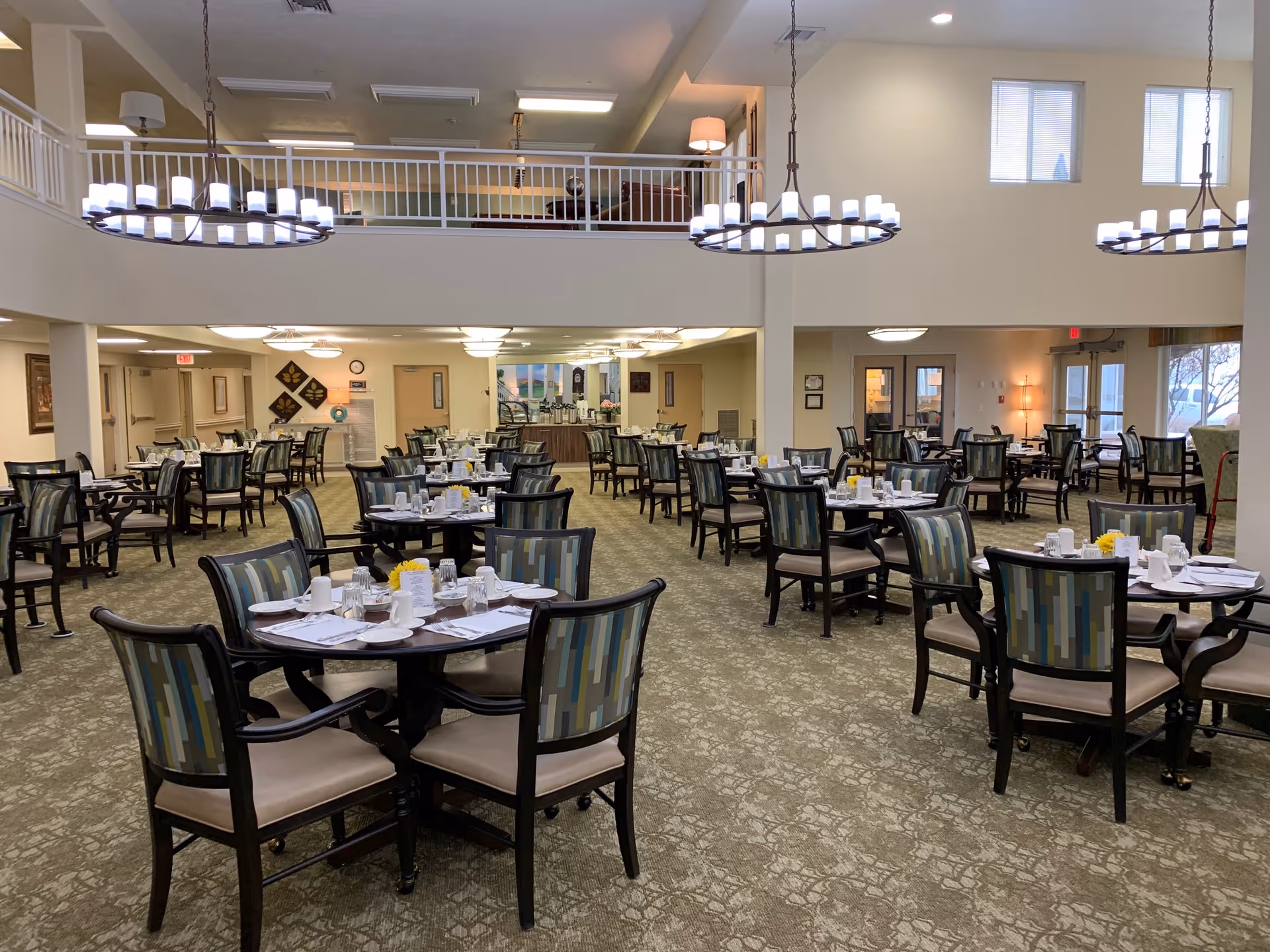 A spacious dining room with multiple round tables set for a meal. Each table has four chairs with patterned upholstery, white plates, cups, and napkins neatly arranged. The room features high ceilings with large circular chandeliers, carpeted floors, and a balcony area above. Large windows allow natural light to enter the room.