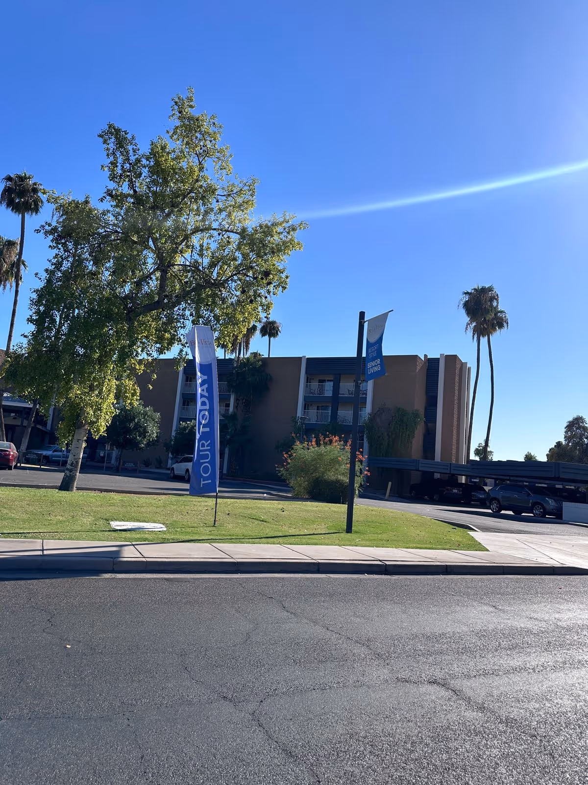 Exterior front view of a senior living building with flags on a grassy lawn, palm trees, and a clear blue sky.
