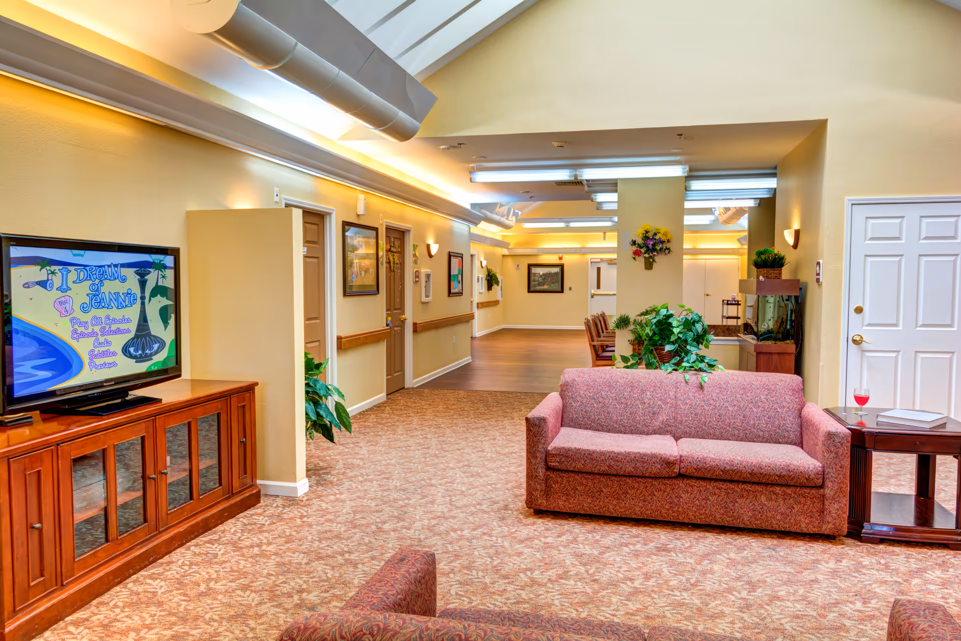A cozy common area in a senior living facility with a patterned carpet, a maroon upholstered sofa, a wooden side table with a glass of red drink and a book, a wooden TV stand with a television displaying a colorful screen, and several framed pictures on the walls. The space is well-lit with ceiling lights and has plants and flowers adding greenery.