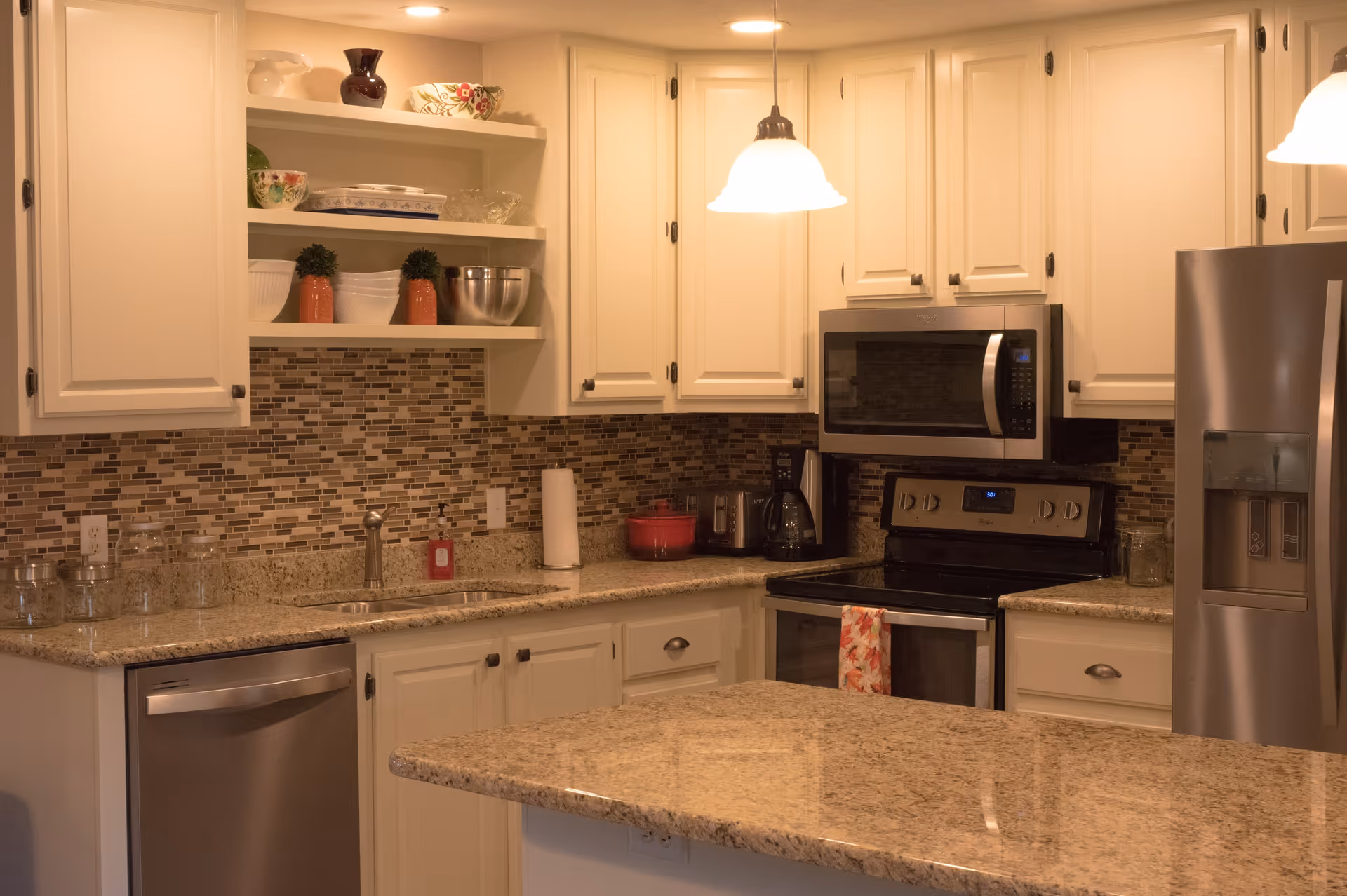 A modern kitchen with beige granite countertops, white cabinets, and a mosaic tile backsplash. The kitchen features stainless steel appliances including a dishwasher, microwave, stove, and refrigerator. There are open shelves with decorative bowls and plants, a paper towel holder, and a hanging light fixture above the counter.