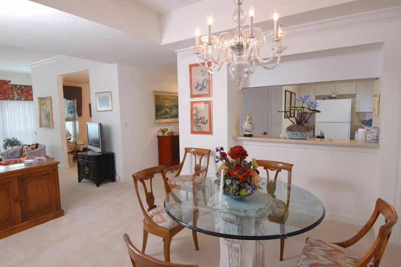 Glass-top round dining table with a floral centerpiece and wooden chairs beneath a chandelier, with a kitchen pass-through and living area visible in the background.
