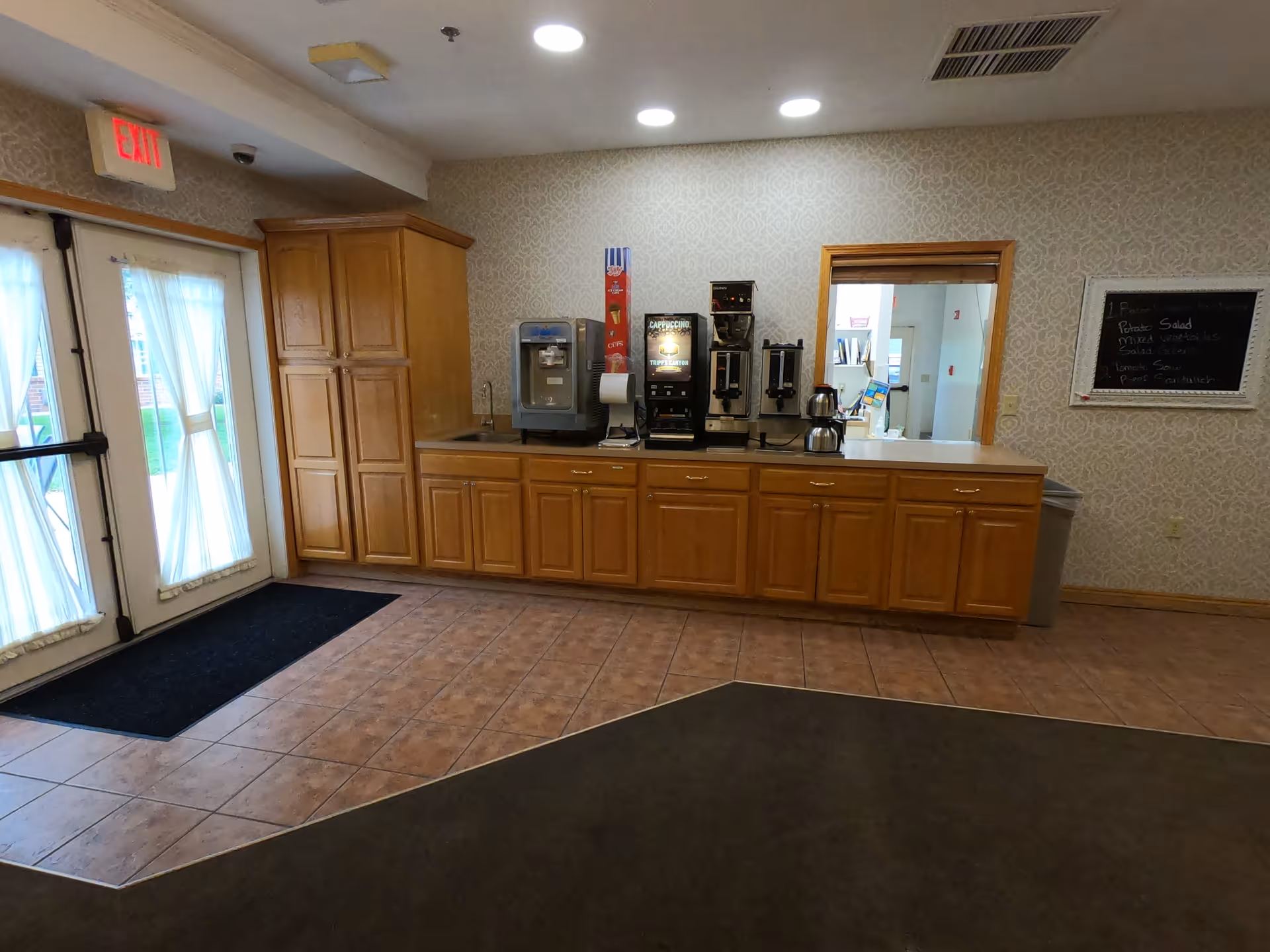 Interior view of a common area with a wooden cabinet counter holding a water dispenser, coffee machines, and a coffee pot. There is a window-like opening above the counter showing another room. To the left, there are double glass doors with curtains and an exit sign above them. The floor is tiled near the counter and doors, with carpet in the foreground. A small chalkboard with handwritten notes is mounted on the wall to the right.