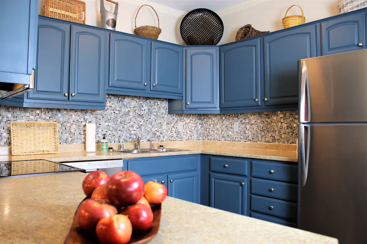 A kitchen with blue cabinets, a mosaic tile backsplash, beige countertops, a stainless steel refrigerator, and a bowl of red apples on the counter. Various wicker baskets are displayed on top of the cabinets.