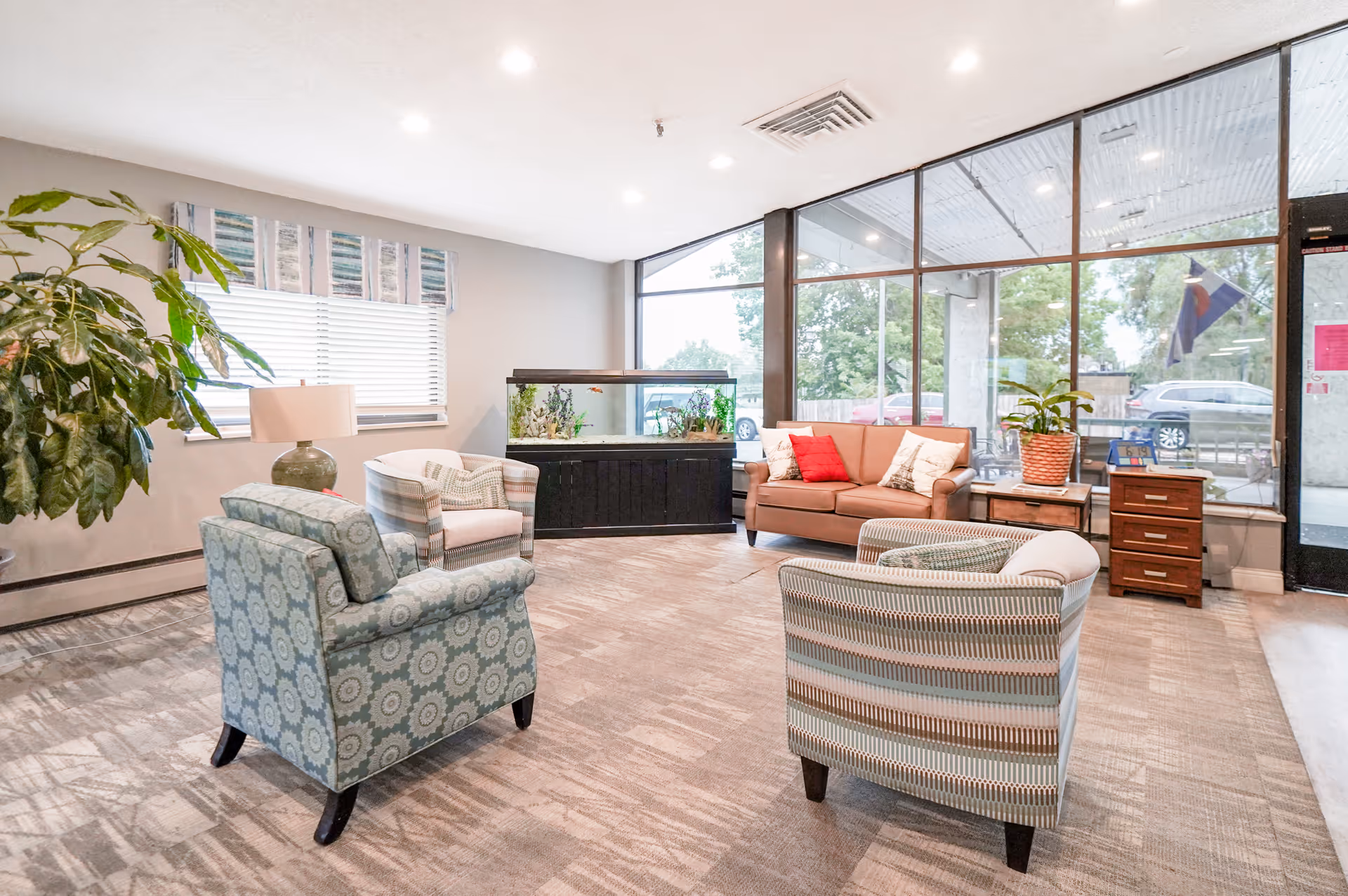A bright and spacious living room area with large windows showing an outside view. The room contains a brown sofa with red and white pillows, three patterned armchairs, a large fish tank on a black stand, a potted plant, a lamp on a side table, and a small wooden drawer unit near the entrance door.