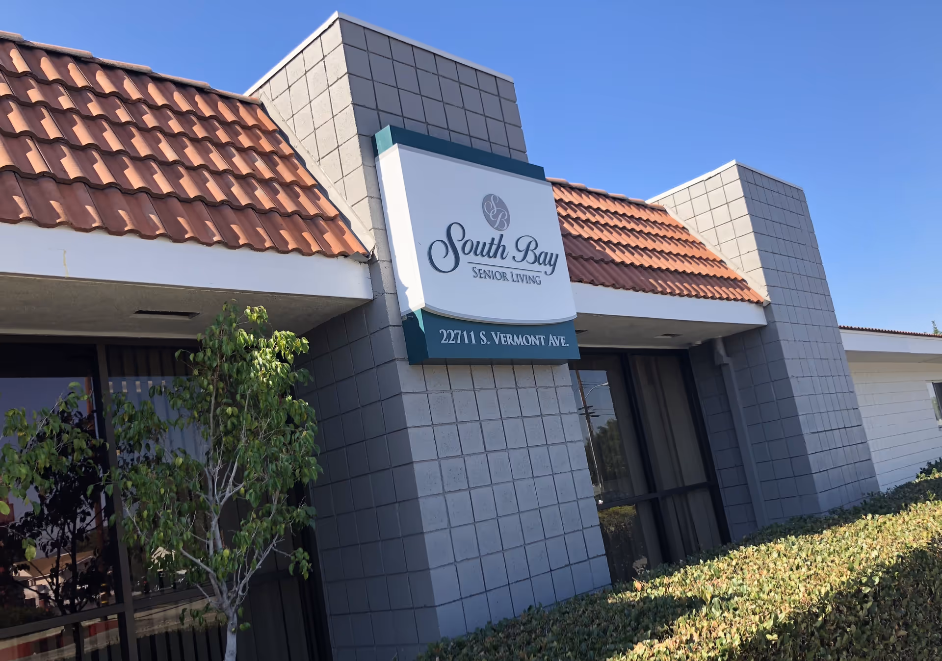 Exterior view of South Bay Senior Living building with a sign displaying the facility name and address 22711 S. Vermont Ave. The building has a tiled roof, gray brick walls, and windows with vertical blinds. There are green bushes and a small tree in front of the building under a clear blue sky.