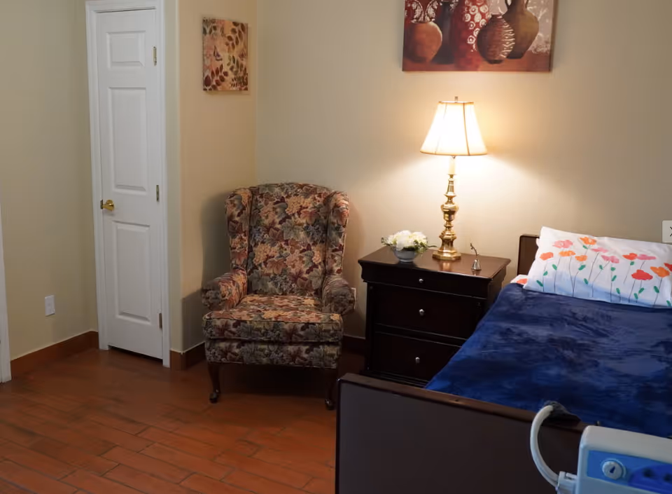 A cozy bedroom corner in an assisted living facility featuring a floral upholstered armchair, a dark wooden nightstand with a brass lamp and a small flower arrangement, and a bed with a blue blanket and a pillow with colorful flower prints. The walls are light beige, and there are two pieces of wall art above the chair and the nightstand. The floor is wooden, and there is a white door with a gold handle on the left side.