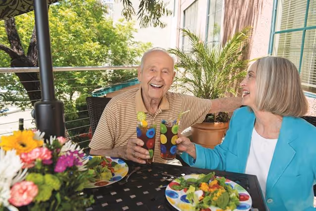 An elderly man and woman sitting at an outdoor table enjoying colorful drinks and salads, smiling and toasting each other on a patio surrounded by greenery.