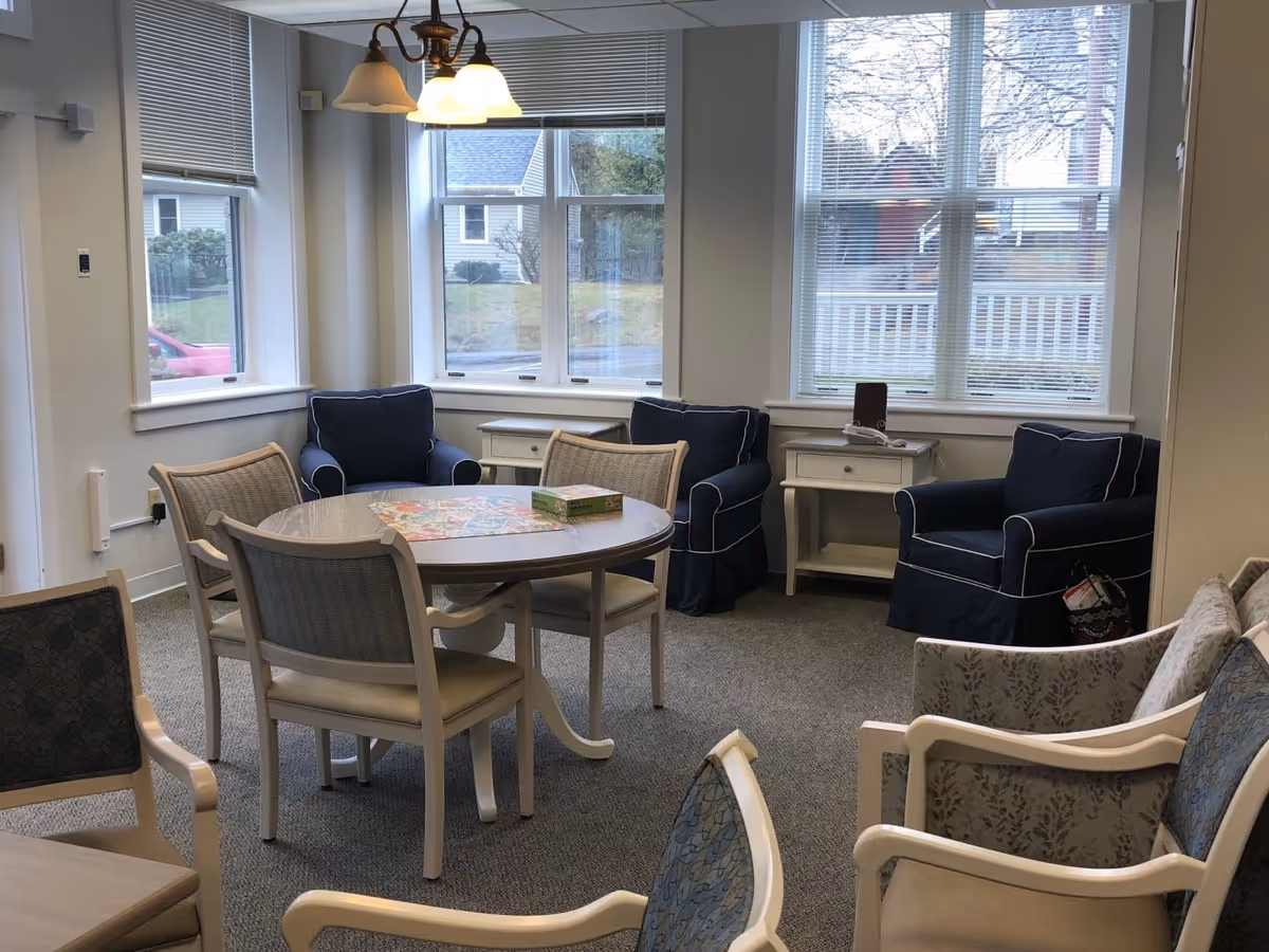 A cozy common area with a round table surrounded by four chairs, two navy blue armchairs with white piping, two white side tables, and additional chairs with patterned upholstery. Large windows with blinds allow natural light to fill the room.
