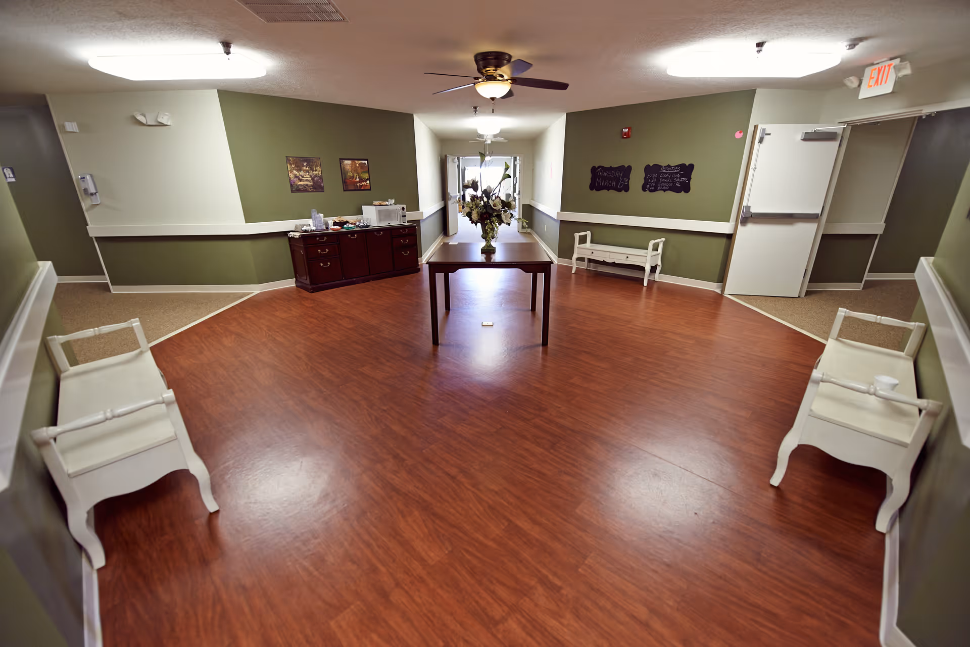 A wide hallway in a nursing home with wood flooring and green walls. There is a wooden table with a flower arrangement in the center, two white benches on either side, a ceiling fan with light, and a sideboard with various items against the wall. The hallway leads to a bright area with glass doors at the far end.