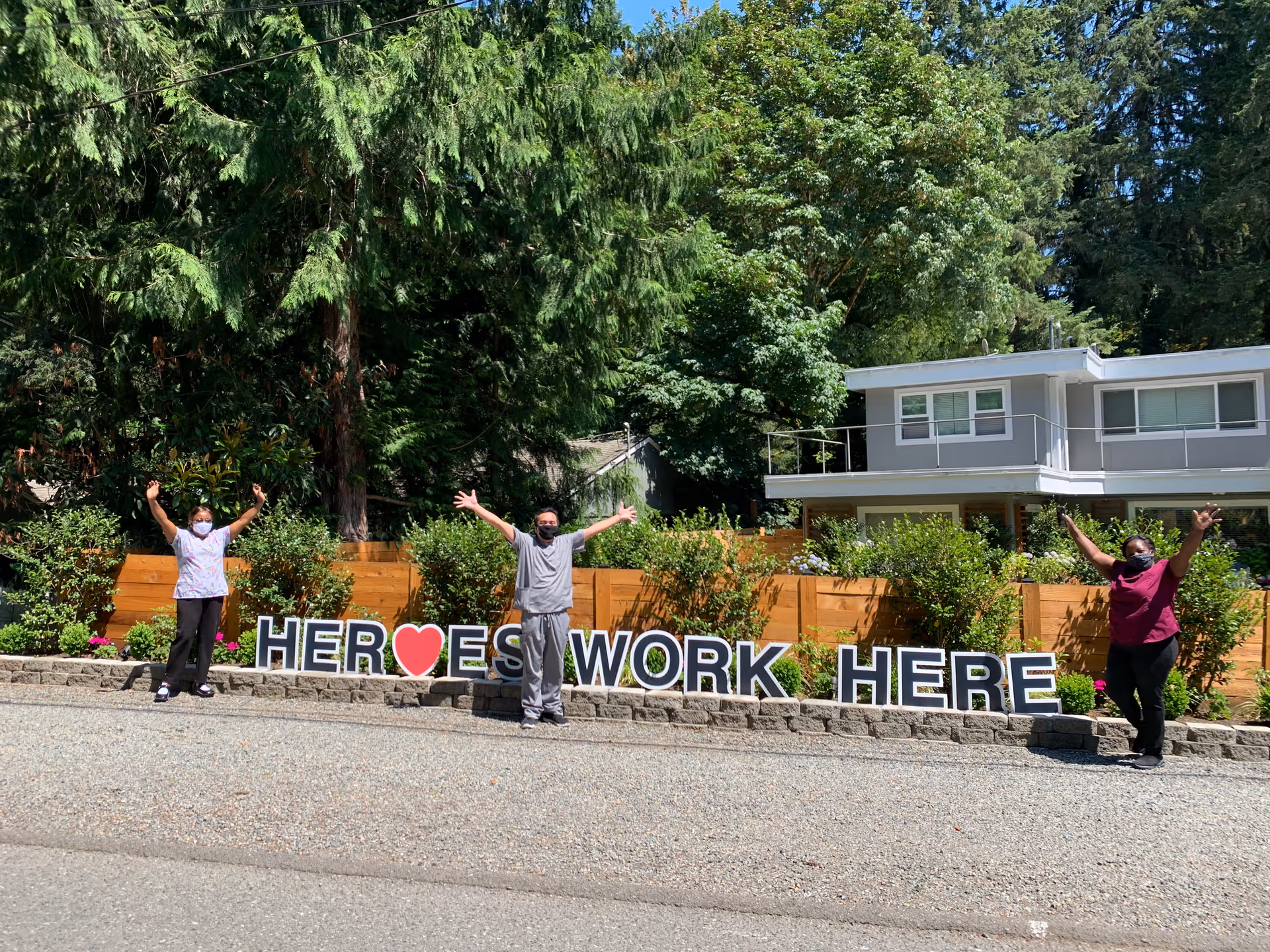Three people wearing masks standing outside in front of a wooden fence and greenery with a sign that reads 'HEROES WORK HERE' with a red heart symbol in place of the letter O in HEROES. They have their arms raised in celebration.