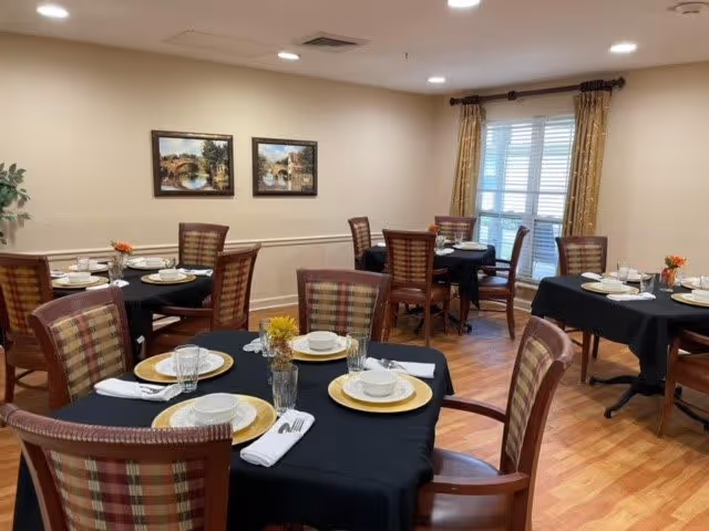 A dining room with several round tables covered with black tablecloths, each set with white plates, bowls, glasses, silverware, and napkins. The chairs have wooden frames with plaid upholstery. The room has light-colored walls with two framed landscape paintings, a window with curtains, and a wooden floor.