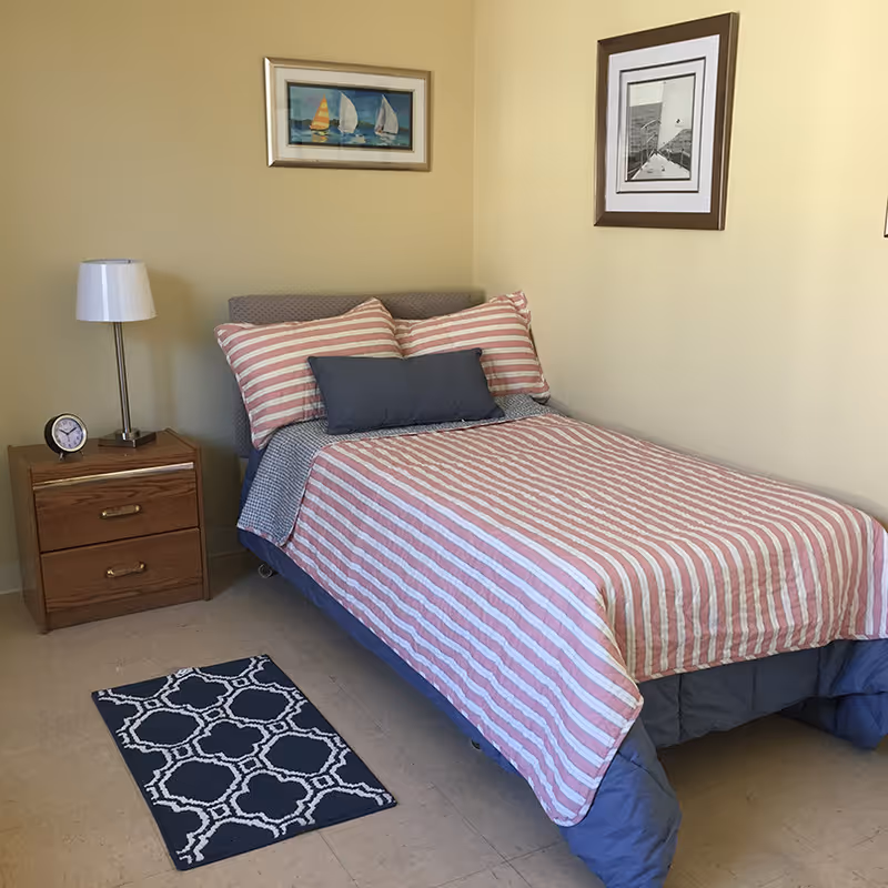 A neatly made single bed with a pink and white striped bedspread and matching pillows, along with a gray lumbar pillow. Next to the bed is a wooden nightstand with two drawers, a small clock, and a white lamp. Two framed pictures hang on the beige walls above the bed. A small dark blue and white patterned rug is on the floor beside the bed.