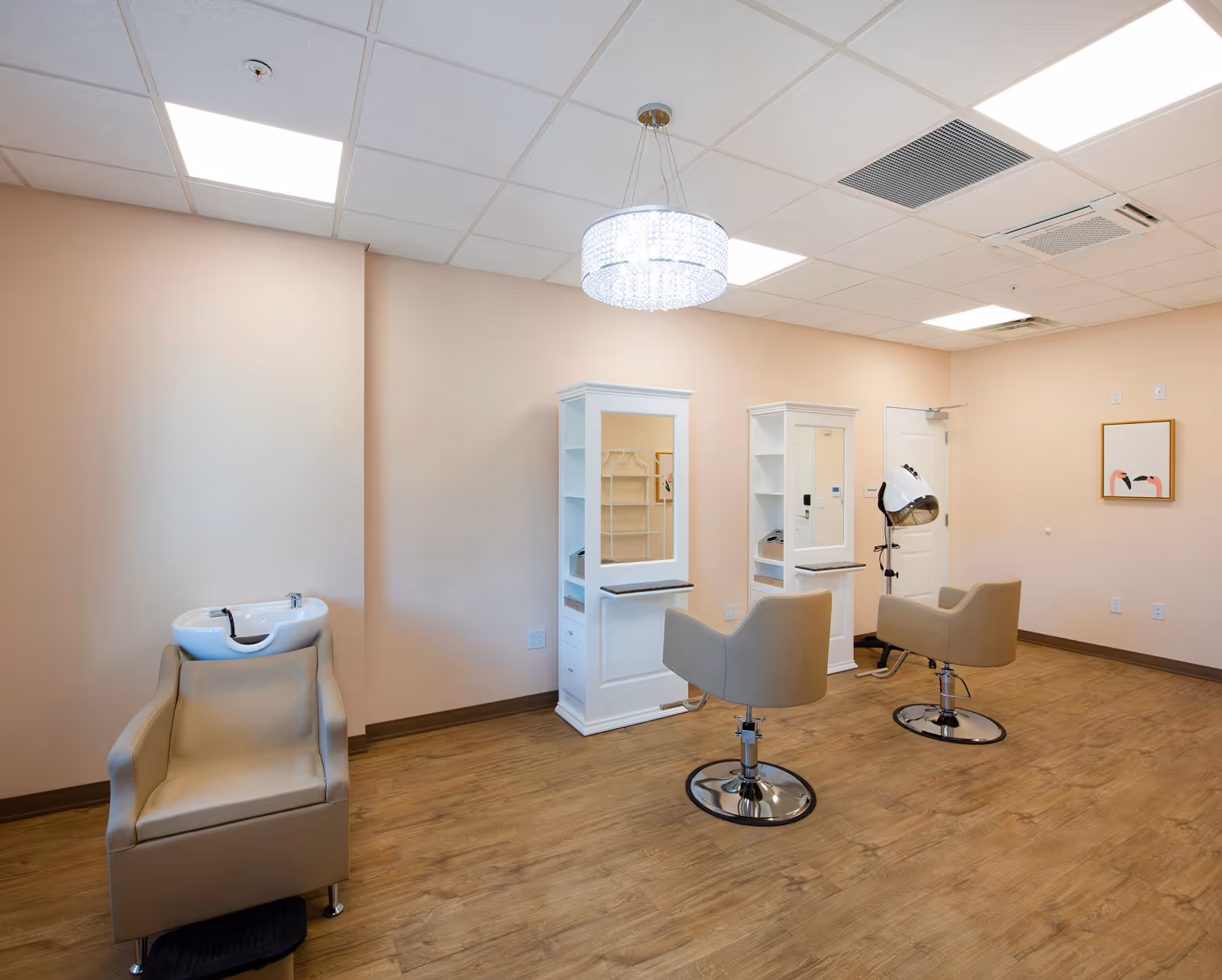 Interior of a salon area in an assisted living facility with two styling stations, each with a mirror, shelf, and beige salon chair. To the left is a beige shampoo chair with a white sink. The room has light peach walls, wood flooring, a white ceiling with recessed lighting, and a modern chandelier hanging from the ceiling. A framed picture is on the far wall.