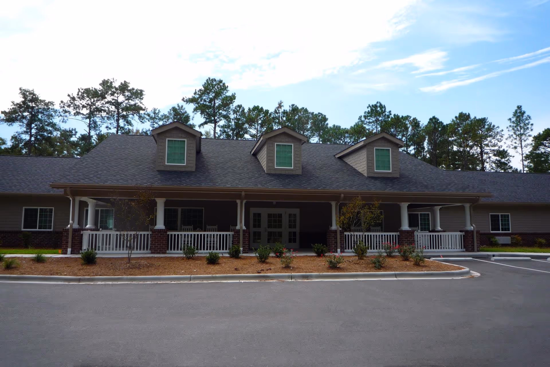 Front exterior view of a single-story building with a dark shingled roof, three dormer windows, a covered porch with white railings and columns, and a landscaped area with small bushes and trees in front. The building is surrounded by tall pine trees under a partly cloudy sky.