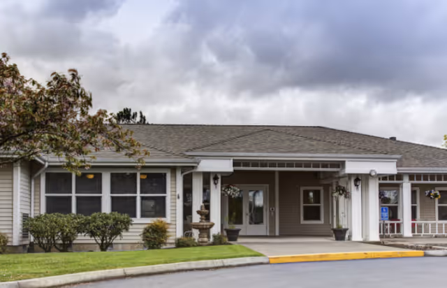 Exterior view of a single-story senior living facility building with beige siding, a covered entrance with white columns, potted plants, and a small fountain near the entrance. There is a tree and shrubs in the landscaped area in front of the building, and a cloudy sky overhead.
