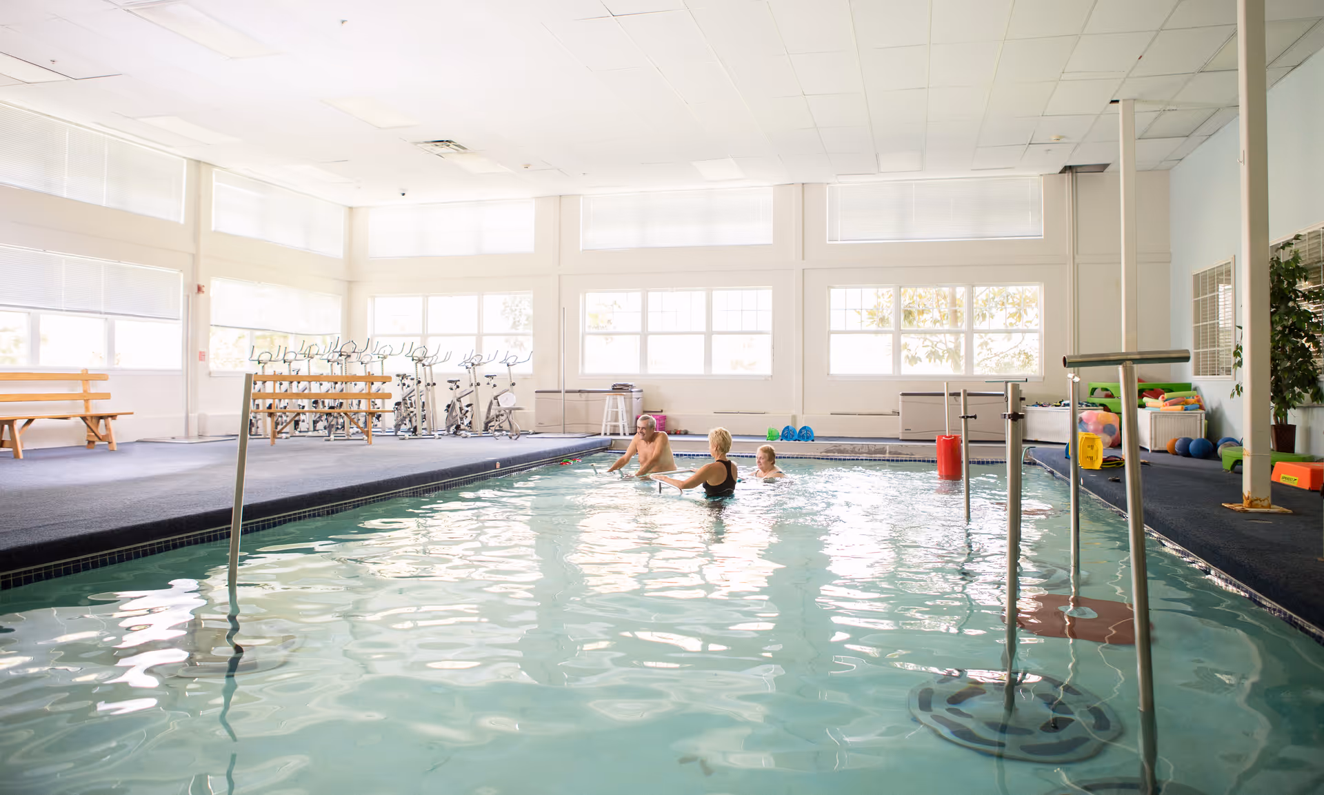 Indoor swimming pool with three people in the water. The pool area is well-lit with large windows and has exercise bikes and benches along the side. Various pool and exercise equipment are visible around the pool area.