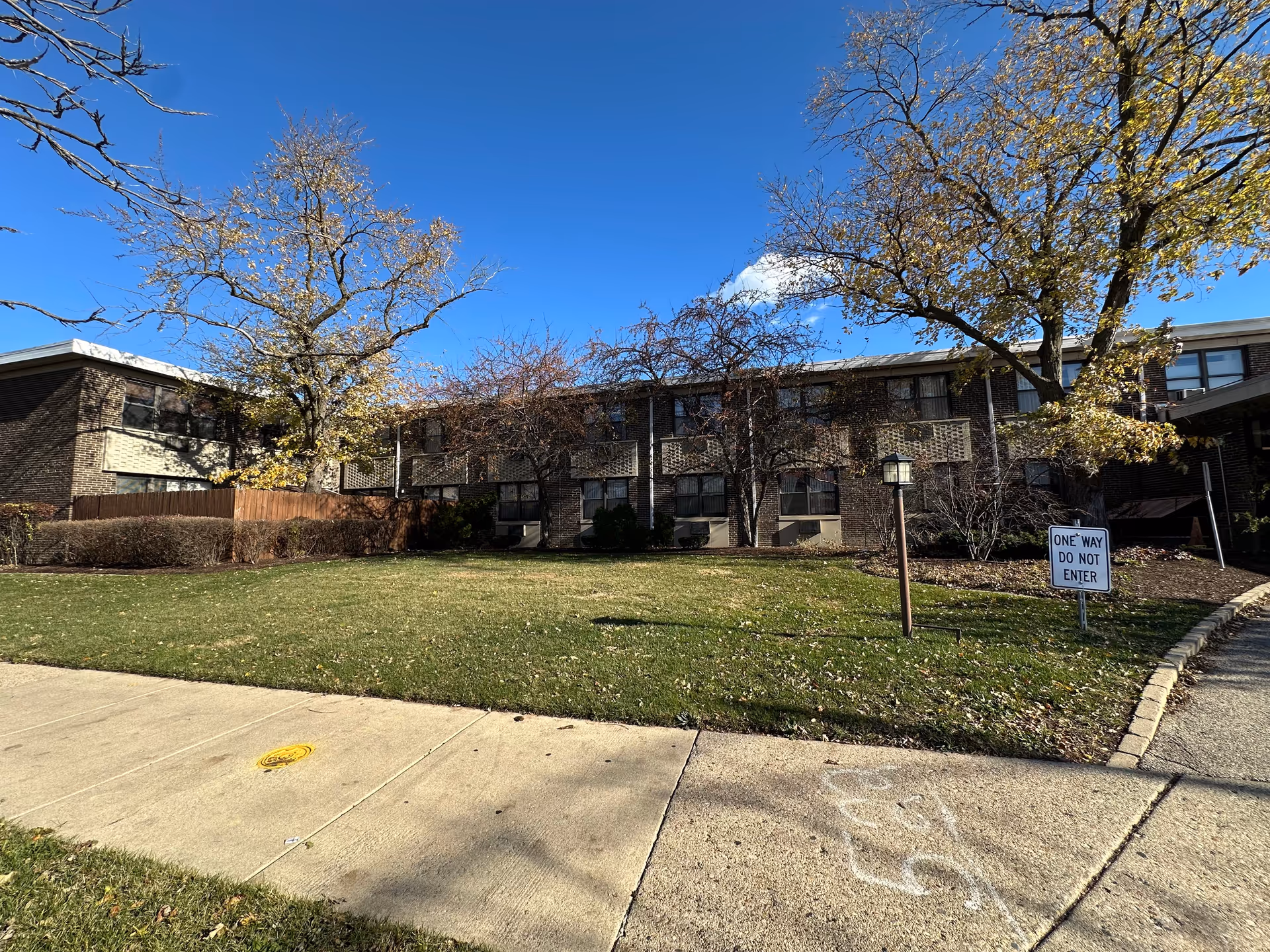 Two-story brick building with a grassy front lawn, trees, sidewalk, and a 'ONE WAY DO NOT ENTER' sign.