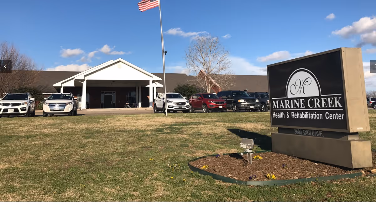 Exterior view of Marine Creek Health & Rehabilitation Center building with a parking lot in front, several parked cars, an American flag on a flagpole, and a large sign displaying the facility's name and address on a grassy area with some flowers.