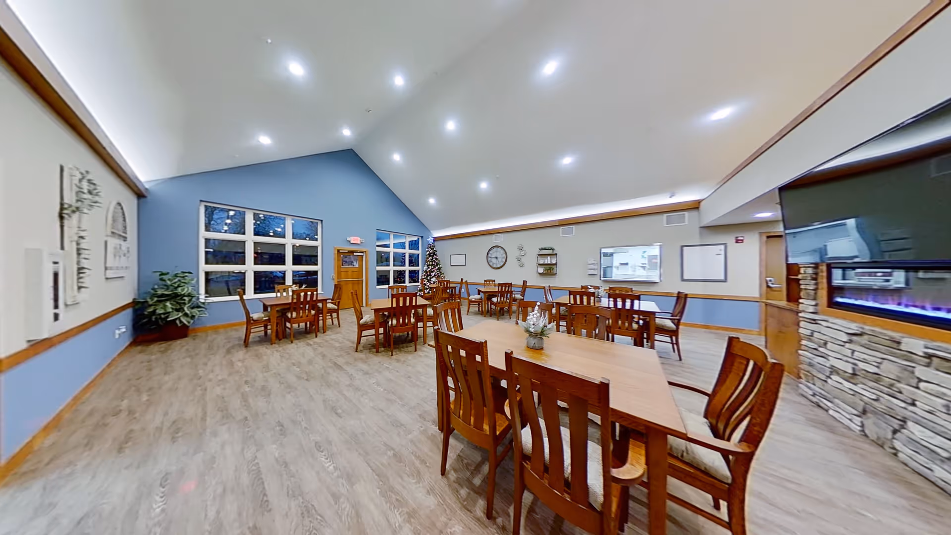 A spacious dining room with multiple wooden tables and chairs arranged neatly. The room features a high vaulted ceiling with recessed lighting, large windows on one side, a decorated Christmas tree in the corner, a wall clock, and a modern electric fireplace with a stone surround. The walls are painted in a combination of blue and beige tones, and there is a small plant in one corner.