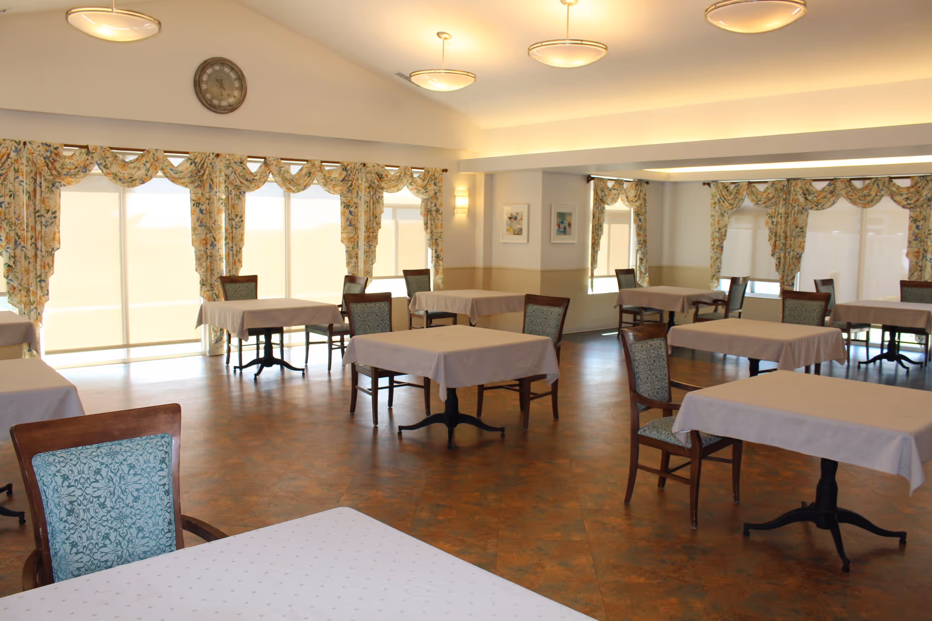 Bright dining room with several tables draped in white tablecloths and wooden chairs, large windows with floral curtains, and overhead lighting.