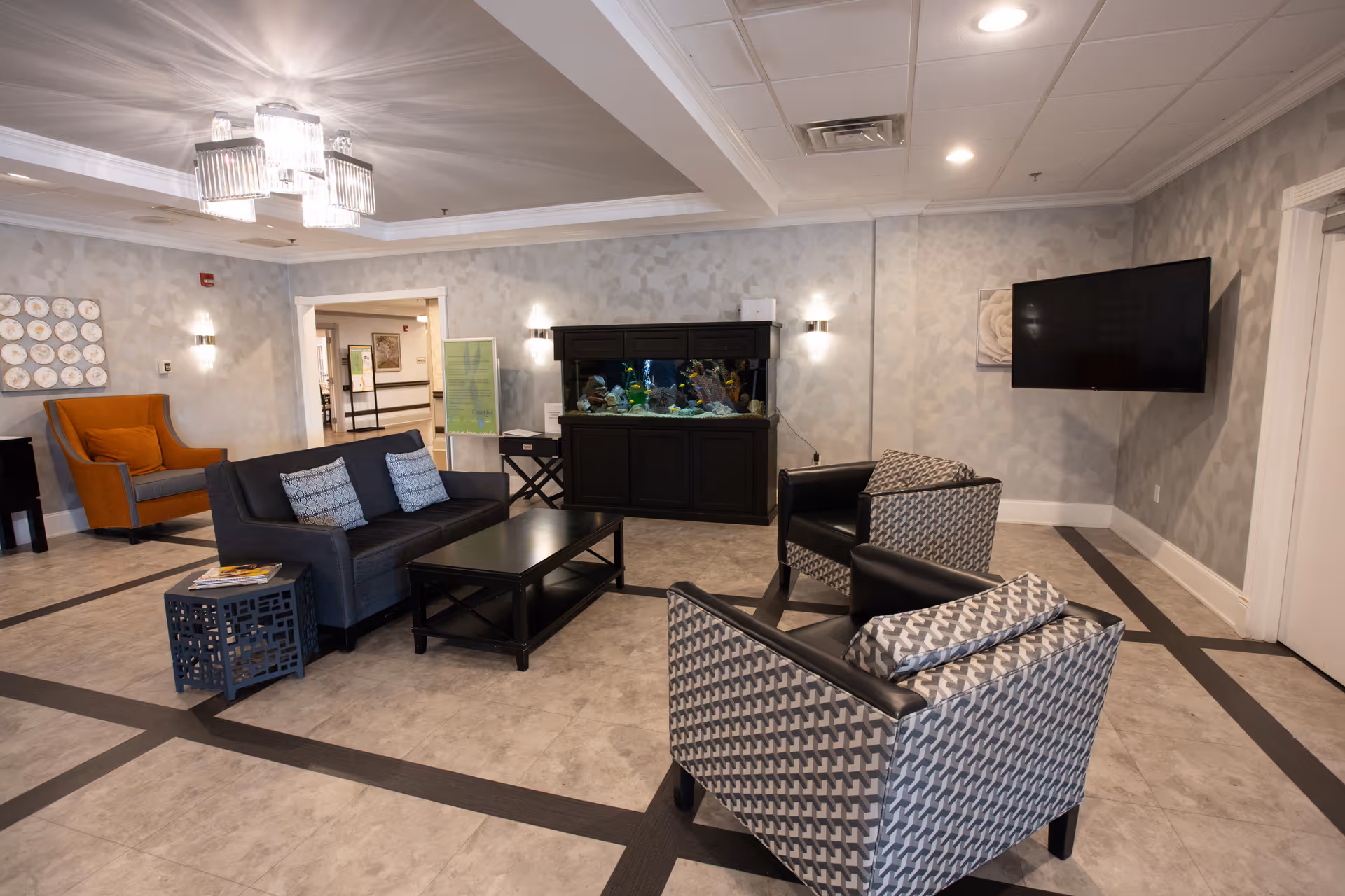 A modern living room area in a senior living facility with a dark gray sofa, two patterned armchairs, a black coffee table, and an orange armchair. There is a large fish tank against the wall, a flat-screen TV mounted on the wall, and decorative wall art. The room has a tiled floor with dark lines and a ceiling light fixture.
