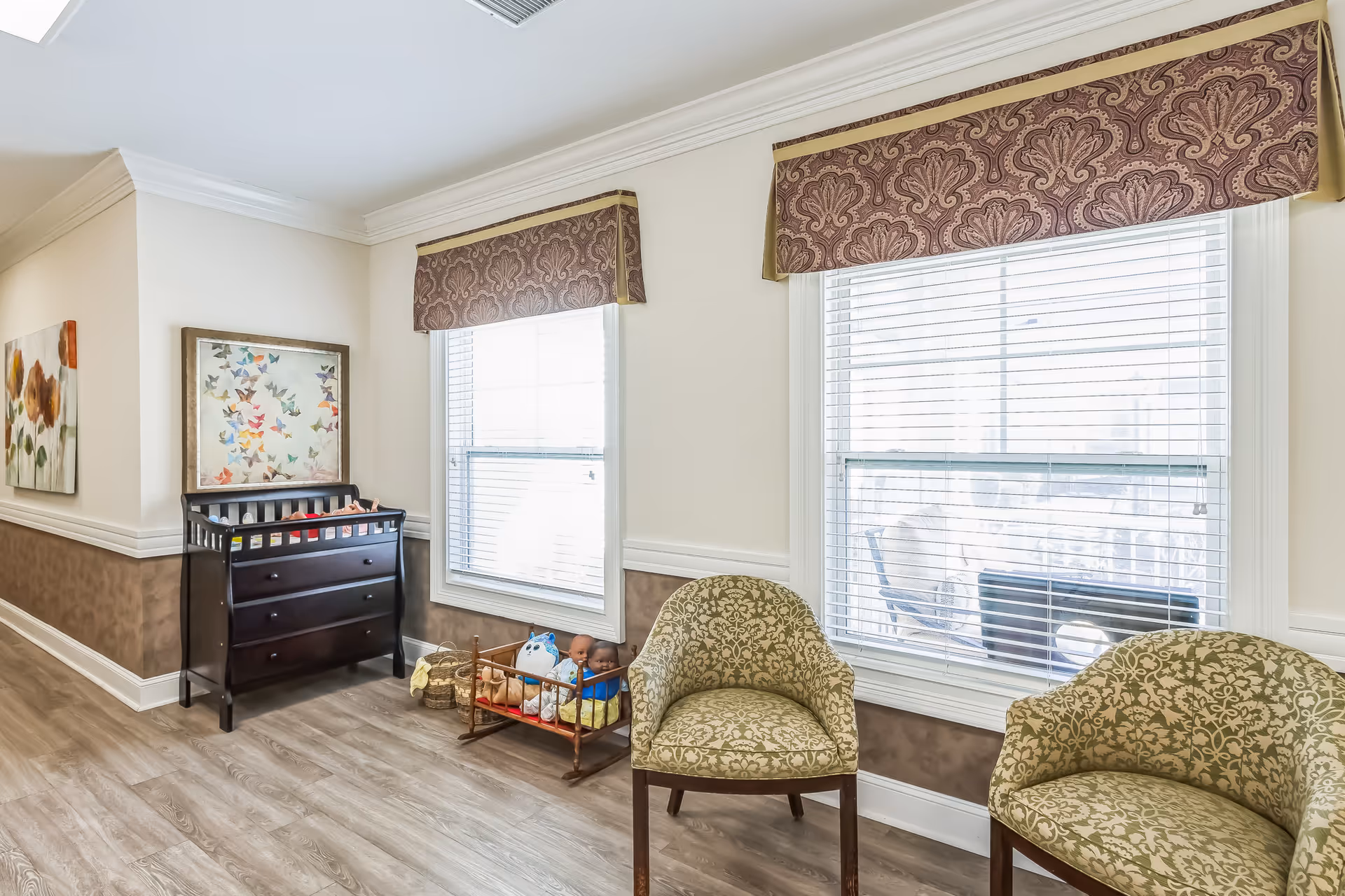 A bright room with two patterned armchairs, a dark wooden dresser with a changing table on top, a basket with toys including a doll and stuffed animals, two large windows with blinds and decorative valances, and framed artwork on the walls.