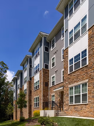 Exterior view of a multi-story residential building with stone and siding facade, many windows, and a small stepped entrance under a clear blue sky.