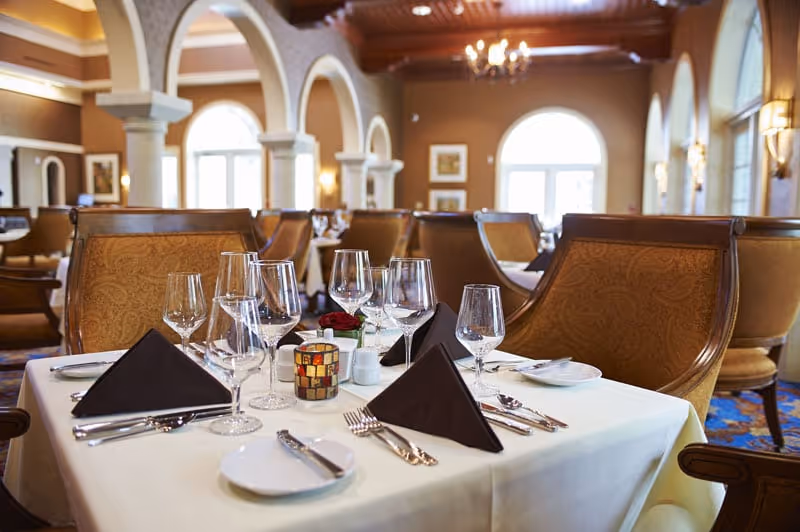Elegant dining room table set with multiple wine glasses, black folded napkins, silverware, and a small decorative candle holder in a spacious room with arched windows and warm lighting.