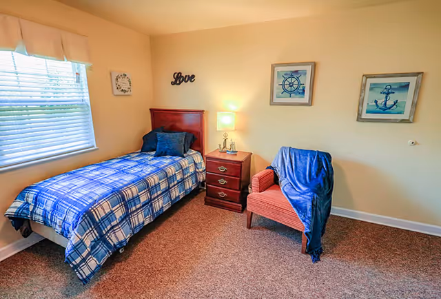 A cozy bedroom with a single bed covered in a blue and white plaid blanket, two dark blue pillows, a wooden headboard, and a nightstand with a lamp and decorative items. There is a red armchair with a blue throw draped over it, beige walls with nautical-themed artwork, and a window with blinds and a valance.