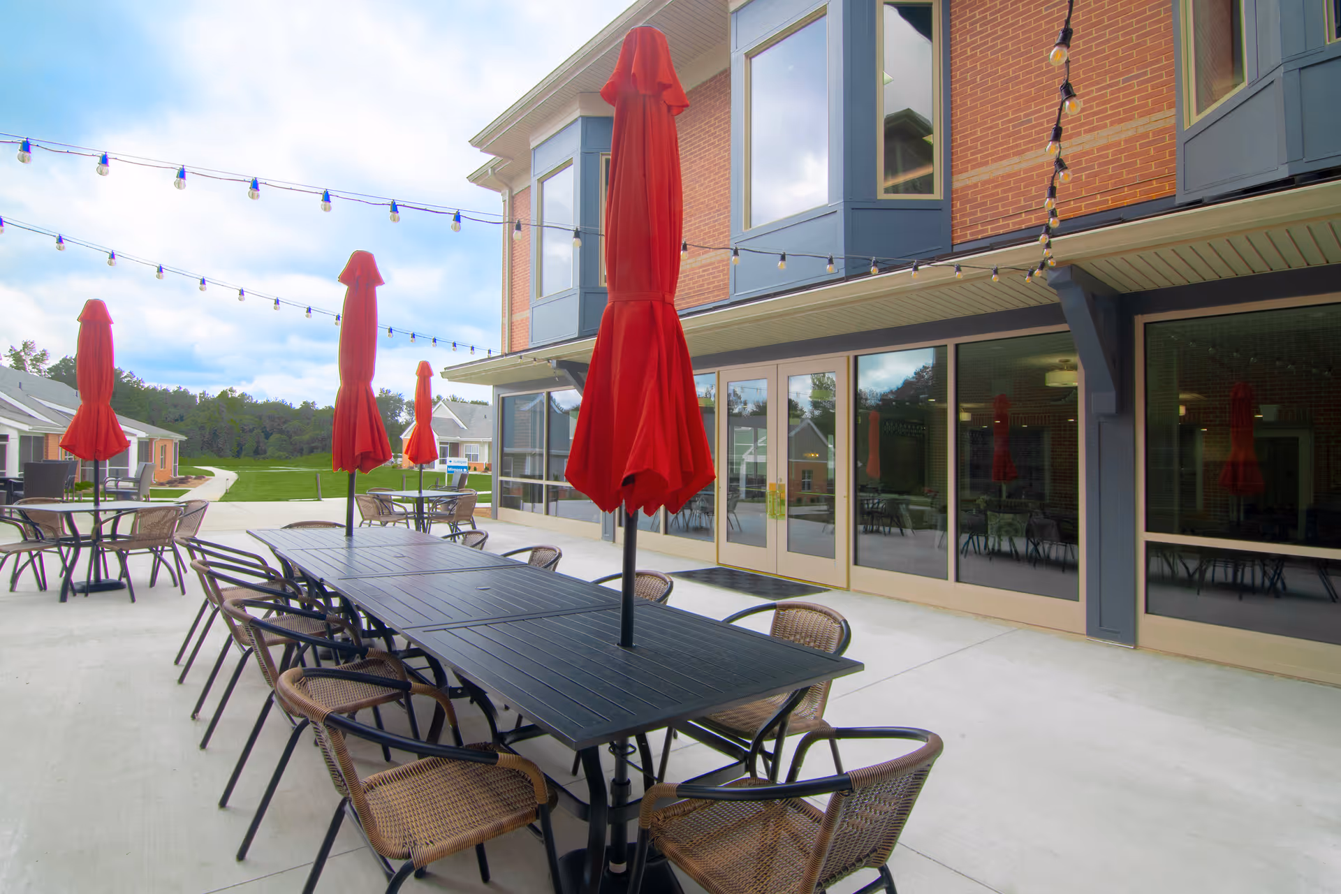 Outdoor patio area at Rosecrest Retirement Community with multiple tables and chairs. Each table has a closed red umbrella, and string lights are hung above the seating area. The patio is adjacent to a brick building with large glass doors and windows.