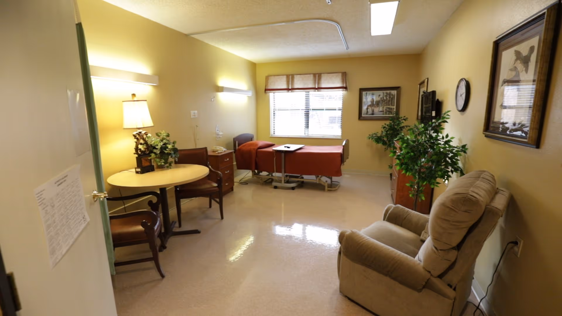 A nursing facility patient room with a hospital bed, recliner, small table and chairs, and decorative plants.