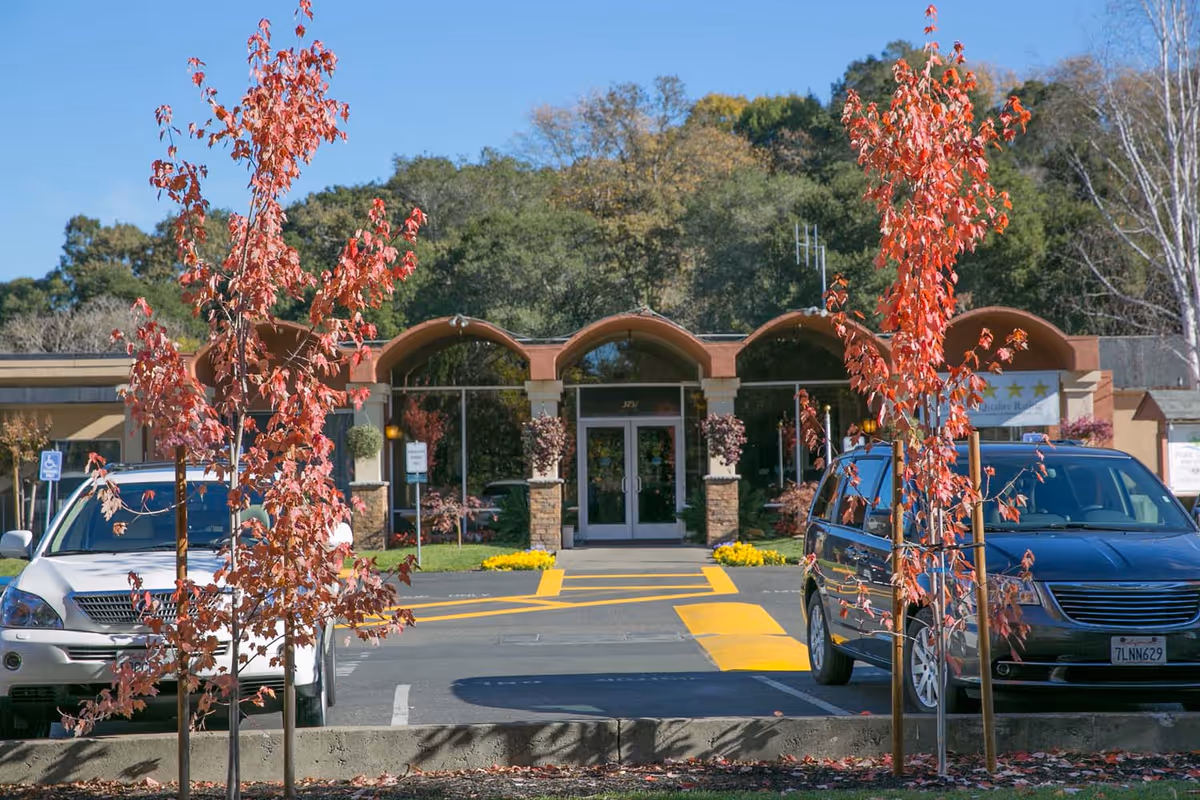 Front exterior view of Park View Post Acute facility with a parking lot in front, two small trees with red leaves, and a building entrance featuring arched roof sections and glass double doors.
