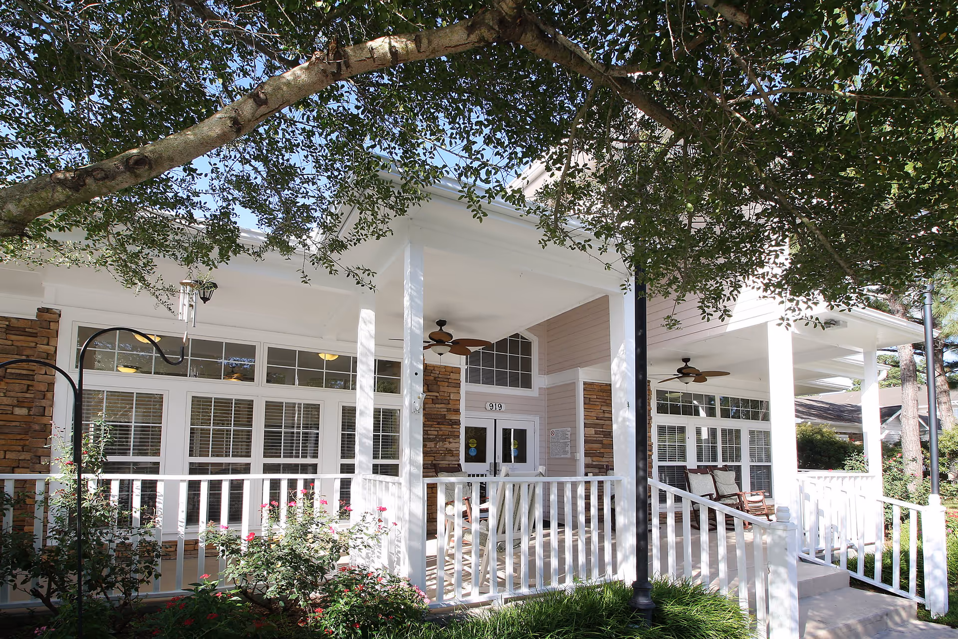 Front porch area of a senior living facility with white railings, rocking chairs, ceiling fans, and large windows. The porch is shaded by a tree with green leaves, and there are flowering bushes in front of the porch.
