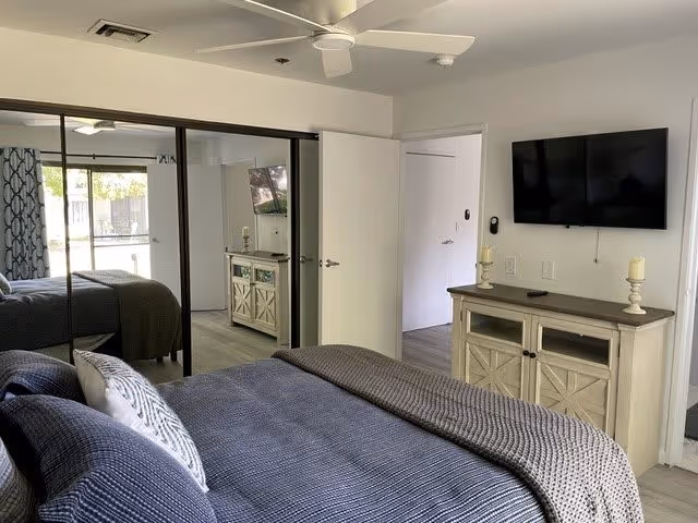 A bedroom in Aasta Assisted Living of Camarillo featuring a neatly made bed with blue and gray bedding, a ceiling fan, a wall-mounted flat screen TV above a wooden cabinet with two candles, mirrored sliding closet doors, and an open door leading to another room with natural light coming through a window.