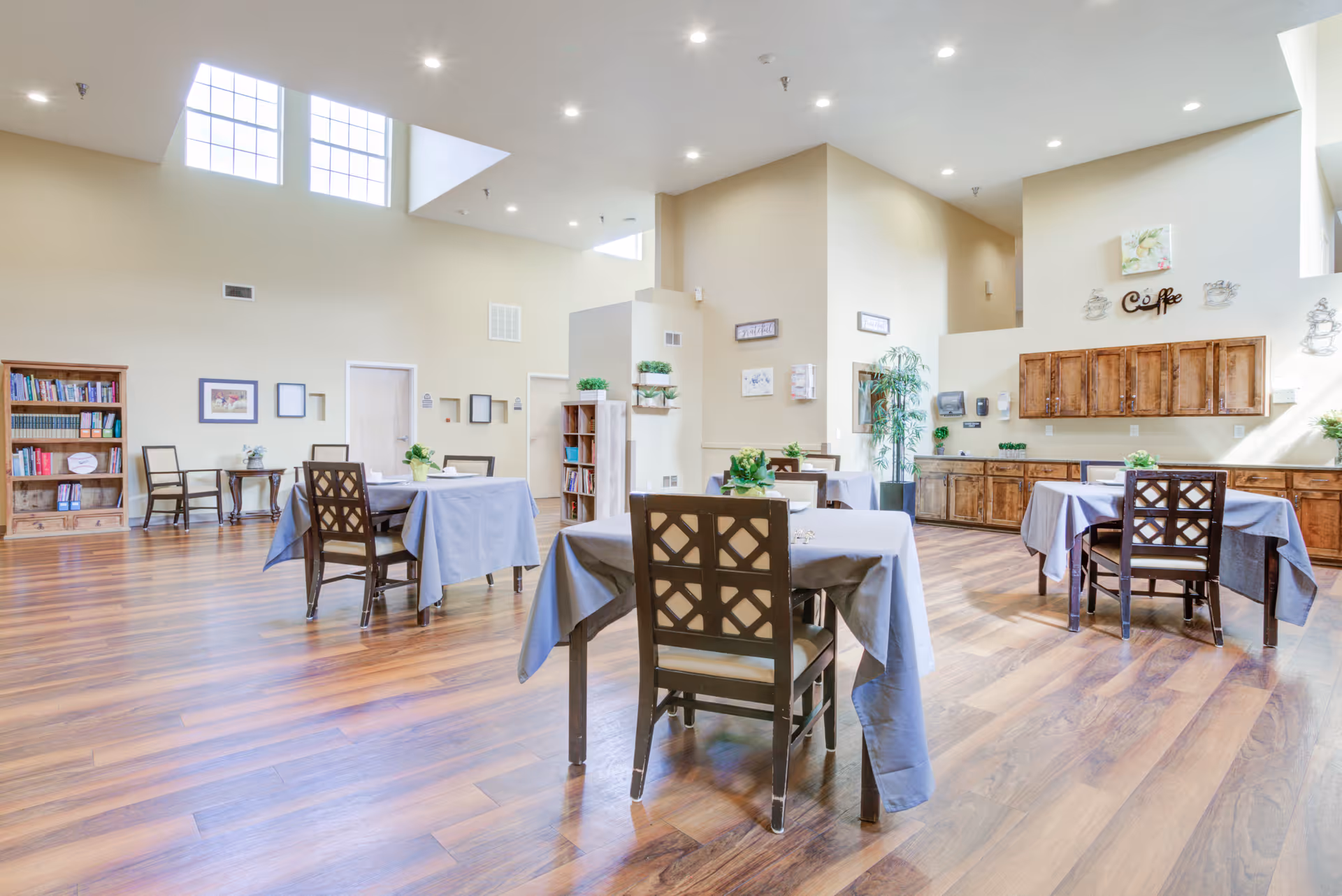 A bright and spacious dining area with several tables covered in gray tablecloths, each surrounded by wooden chairs. The room features high ceilings with large windows allowing natural light to fill the space. There are wooden cabinets along one wall with a decorative 'Coffee' sign above them. Bookshelves and framed pictures decorate the walls, and the floor is covered with wood-patterned flooring.